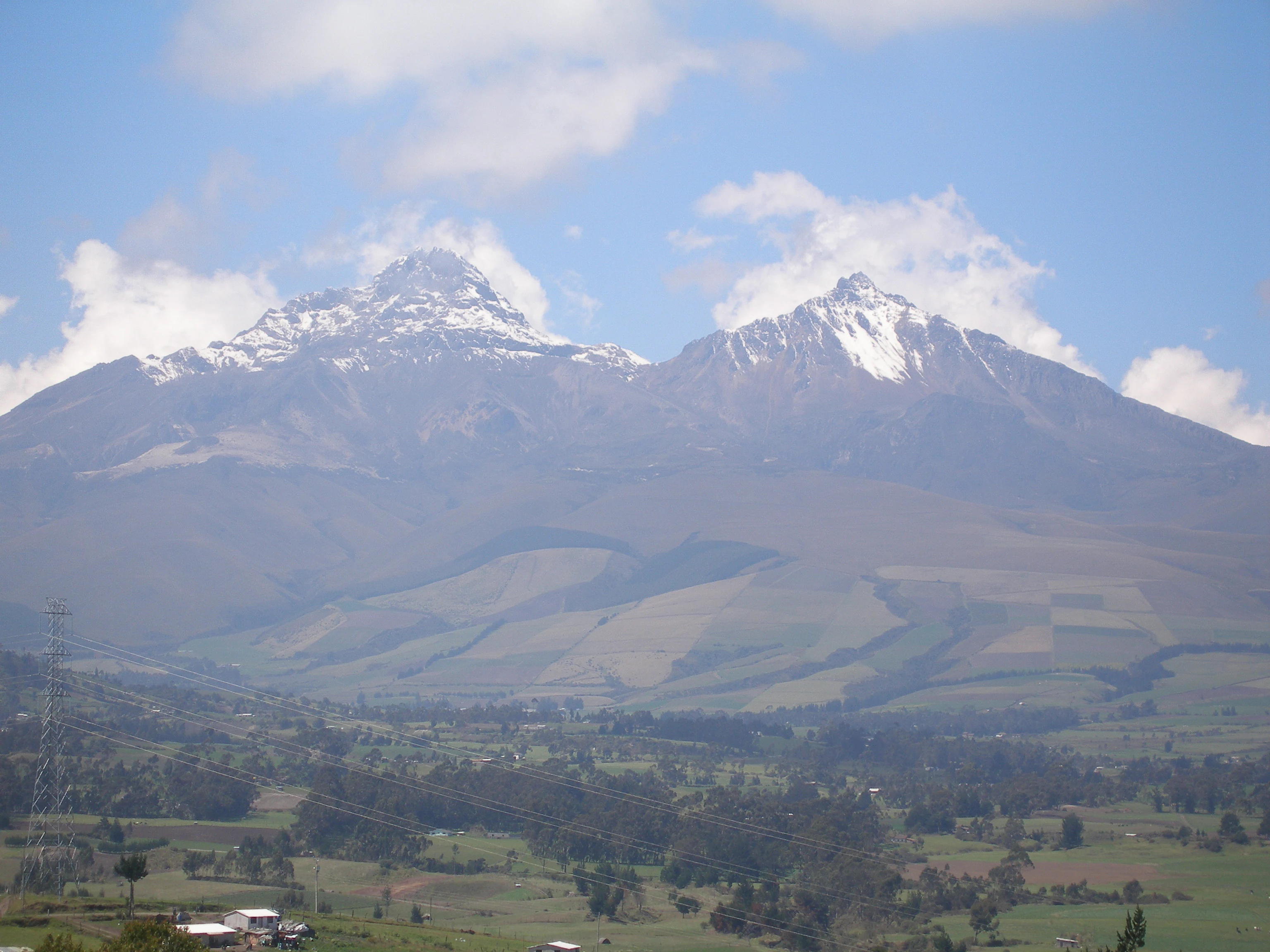 Two large snow-capped mountain peaks rising above a green agricultural valley with scattered trees and buildings under a blue sky with clouds.