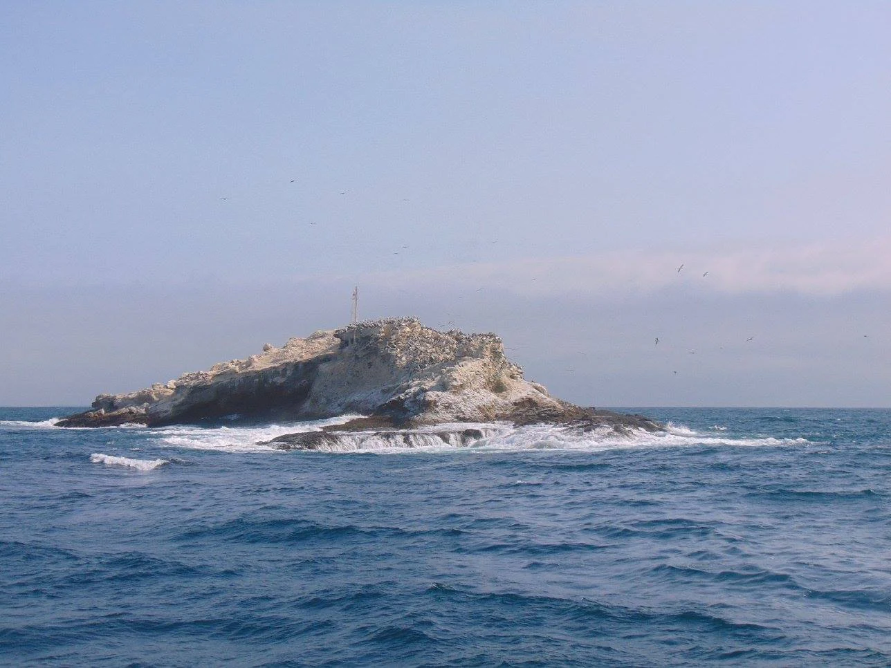 A small, rocky island surrounded by ocean waves under a hazy sky.