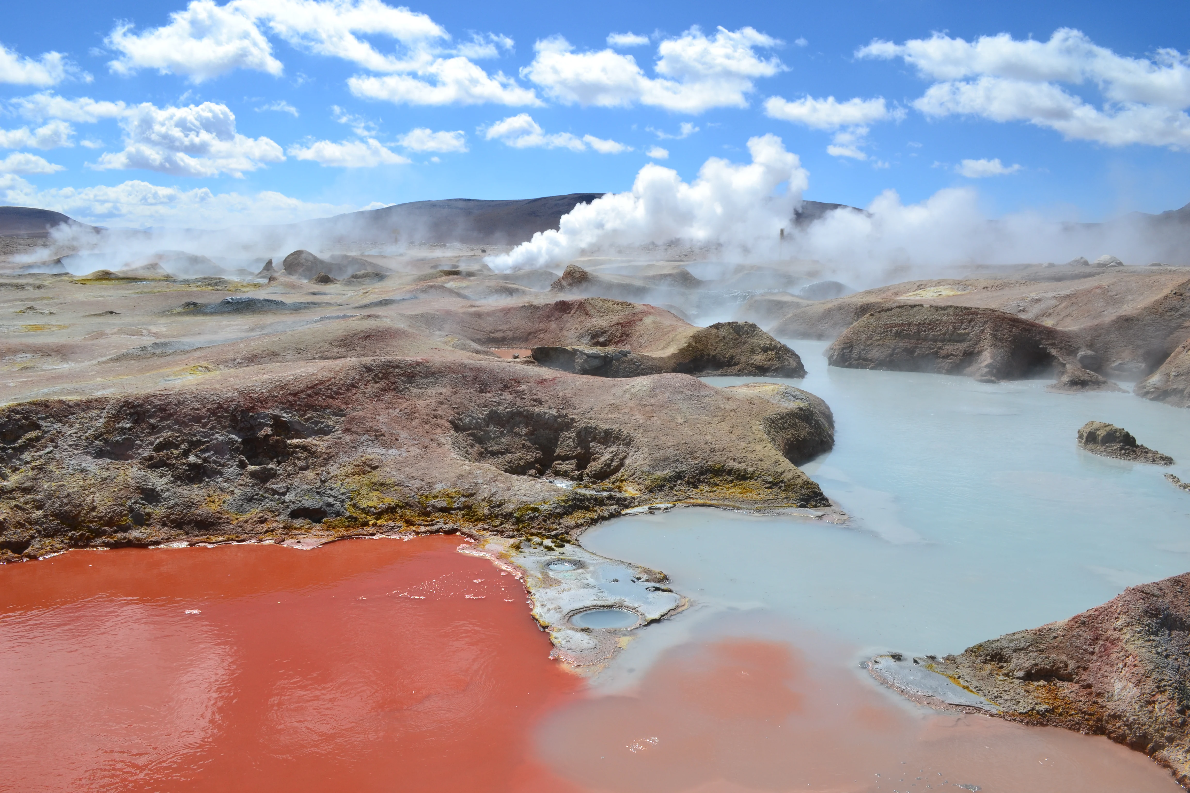 A high-altitude geothermal landscape with a bright red mineral pool in the foreground, adjacent to pools of opaque light-blue water, with steam rising from vents in the barren, rocky terrain under a bright blue sky with clouds.