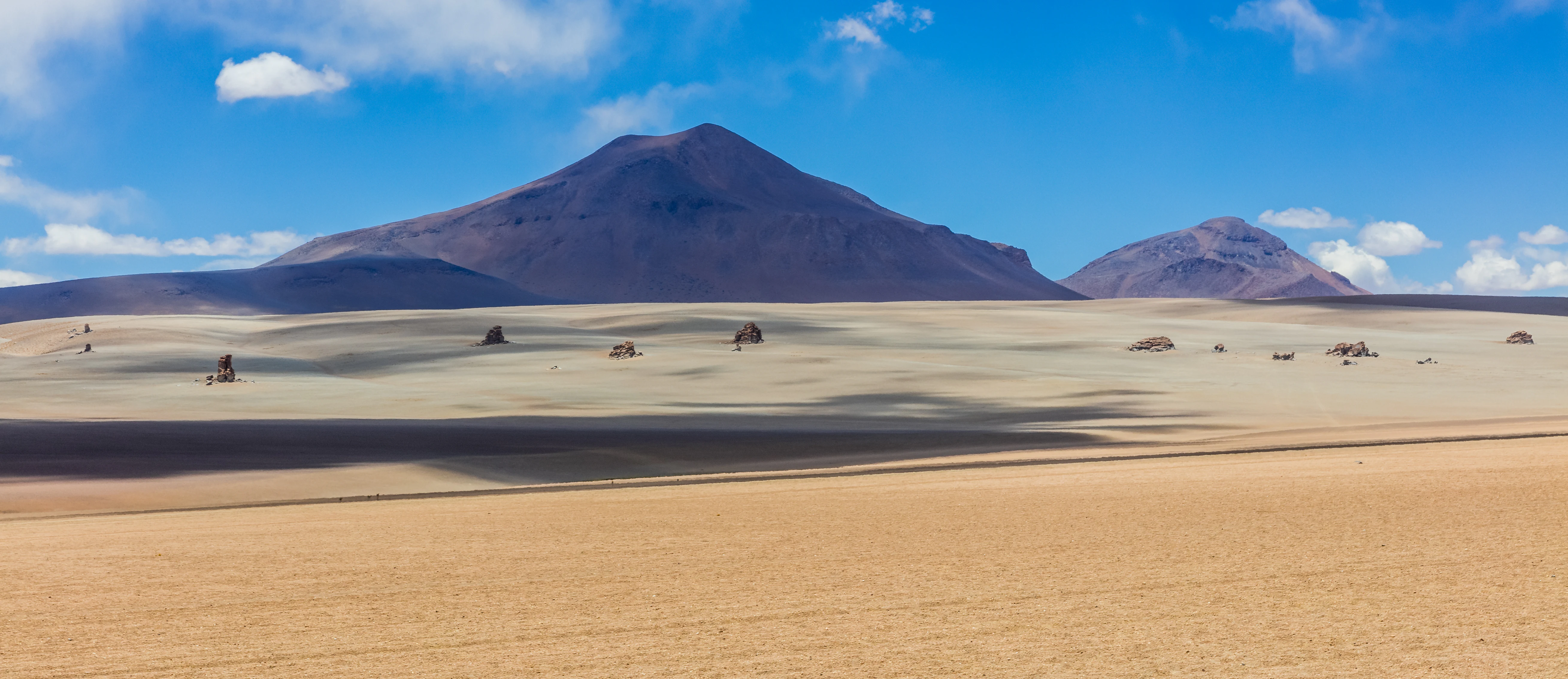 A wide, panoramic view of a vast, beige, sandy desert plain under a blue sky, featuring prominent dark, conical volcanic mountains in the background and sparse, scattered rock formations on the flat terrain.