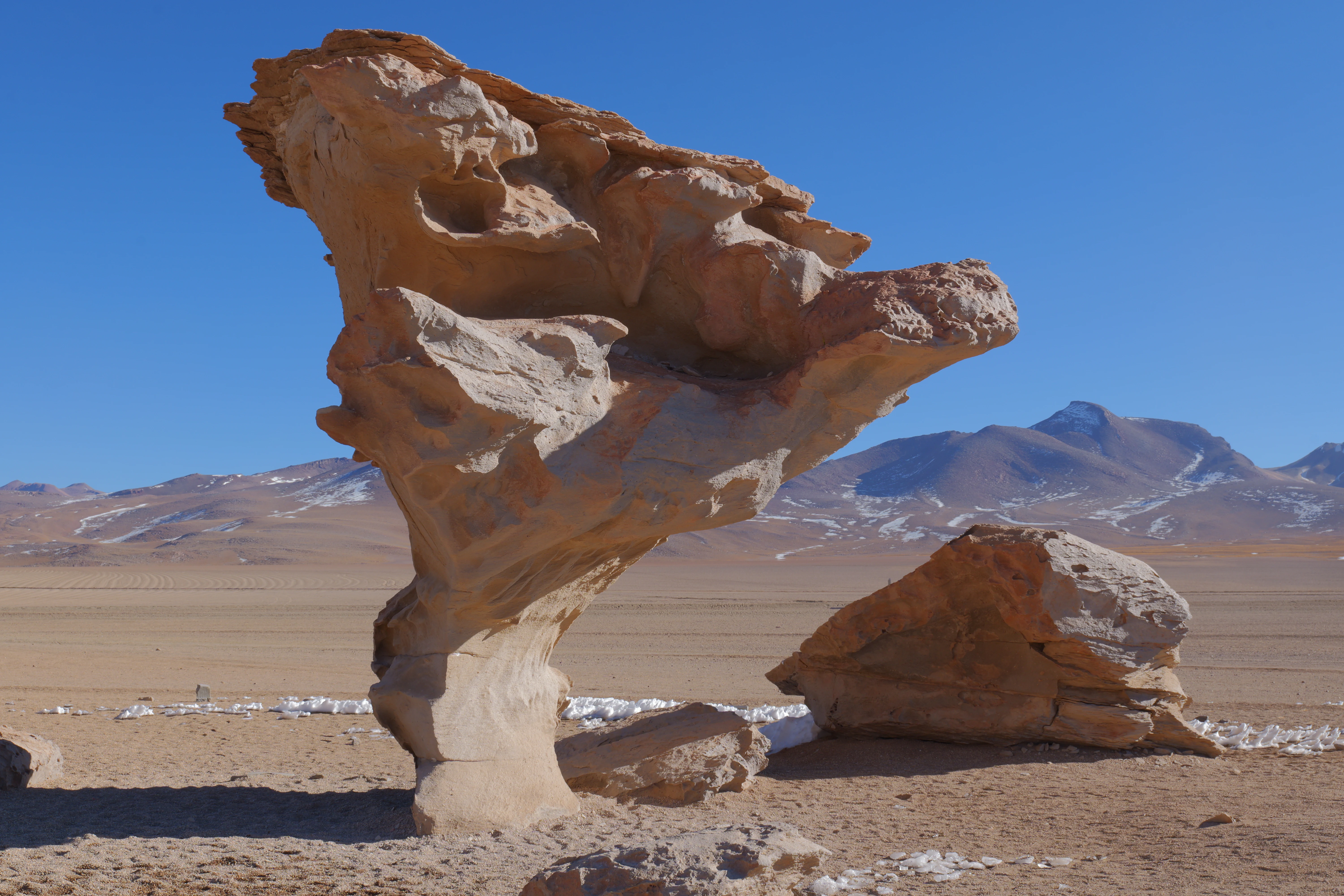 A large, weathered, mushroom-shaped rock formation standing in a flat, arid landscape under a clear blue sky, with a range of distant mountains in the background.
