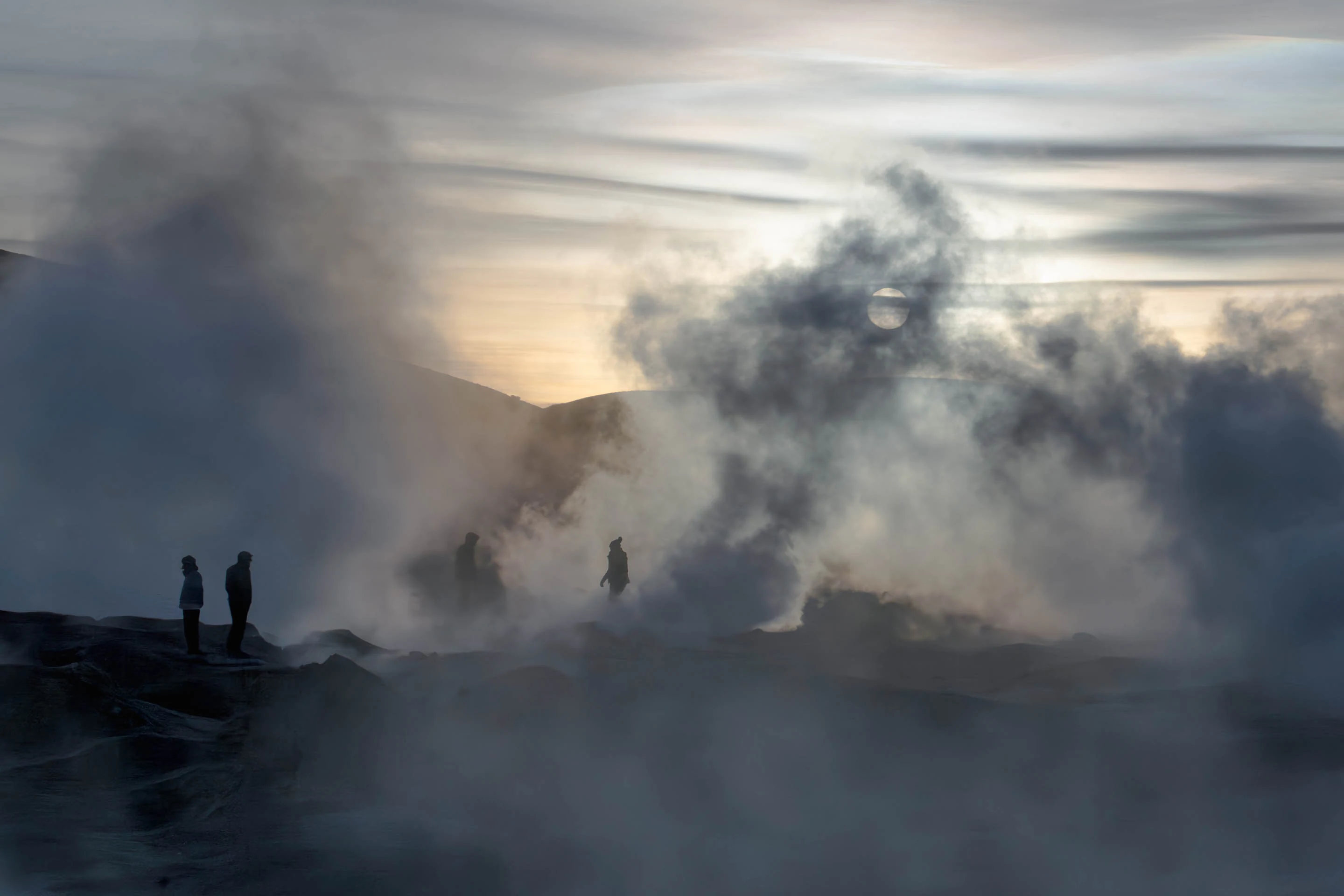 Several people stand near steaming geothermal vents at dawn, with thick plumes of steam obscuring the rocky landscape.