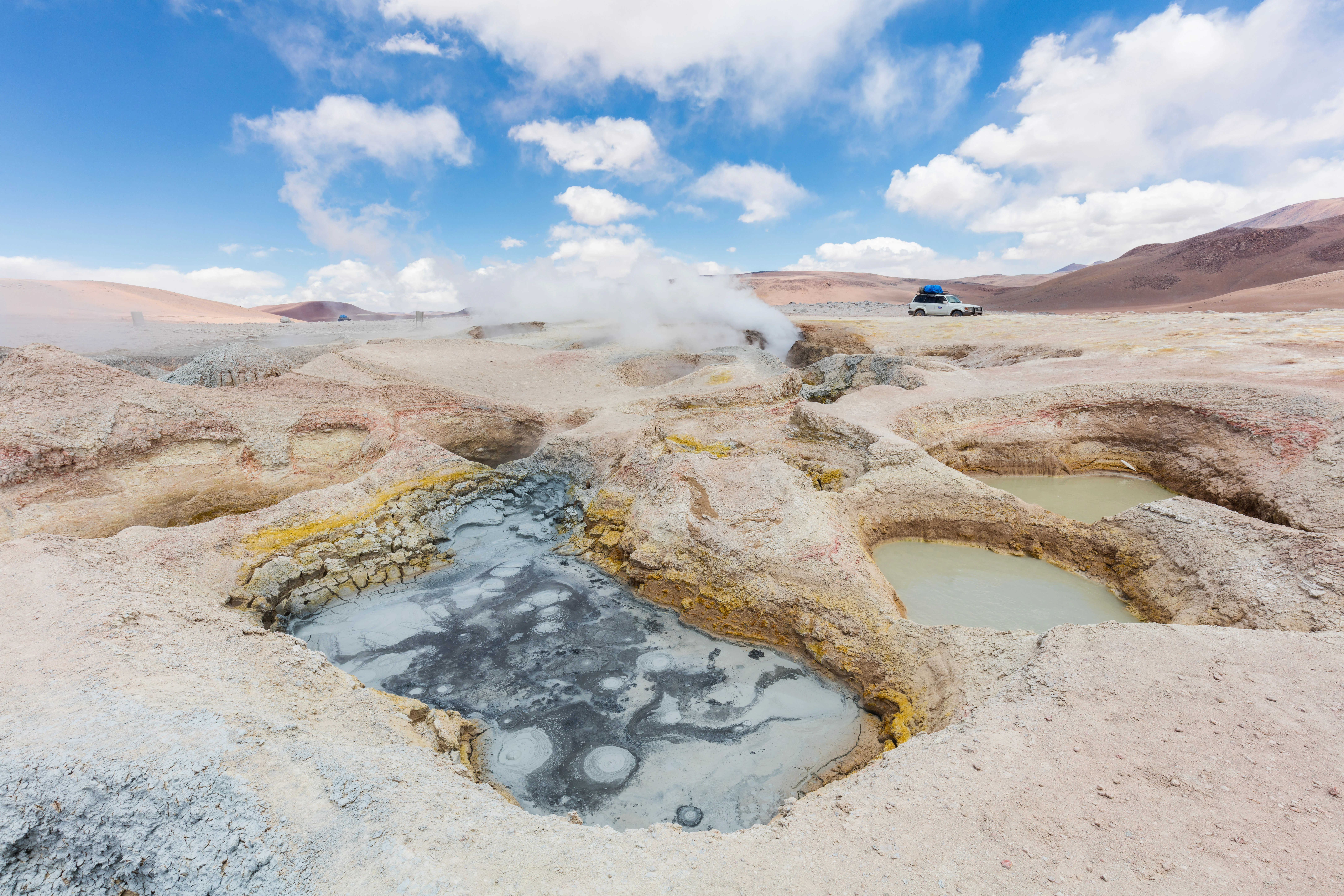 A wide angle landscape view of the Sol de Mañana geyser field with bubbling mud pools in the foreground, steam rising in the middle, and barren volcanic terrain under a blue sky.