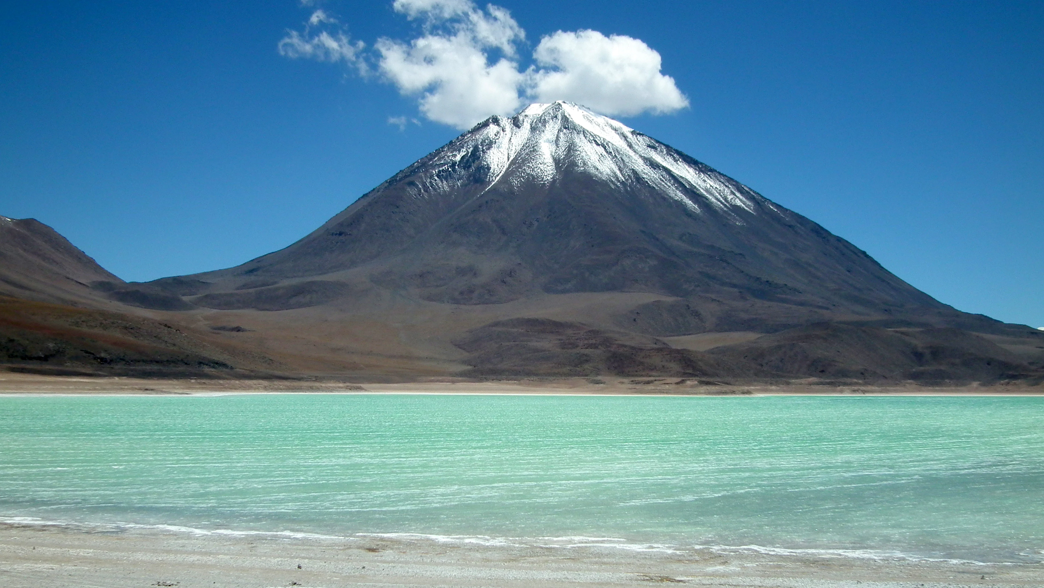 A wide view of a turquoise alpine lake in the foreground with a prominent, conical, snow-capped volcano rising sharply in the background under a clear blue sky.