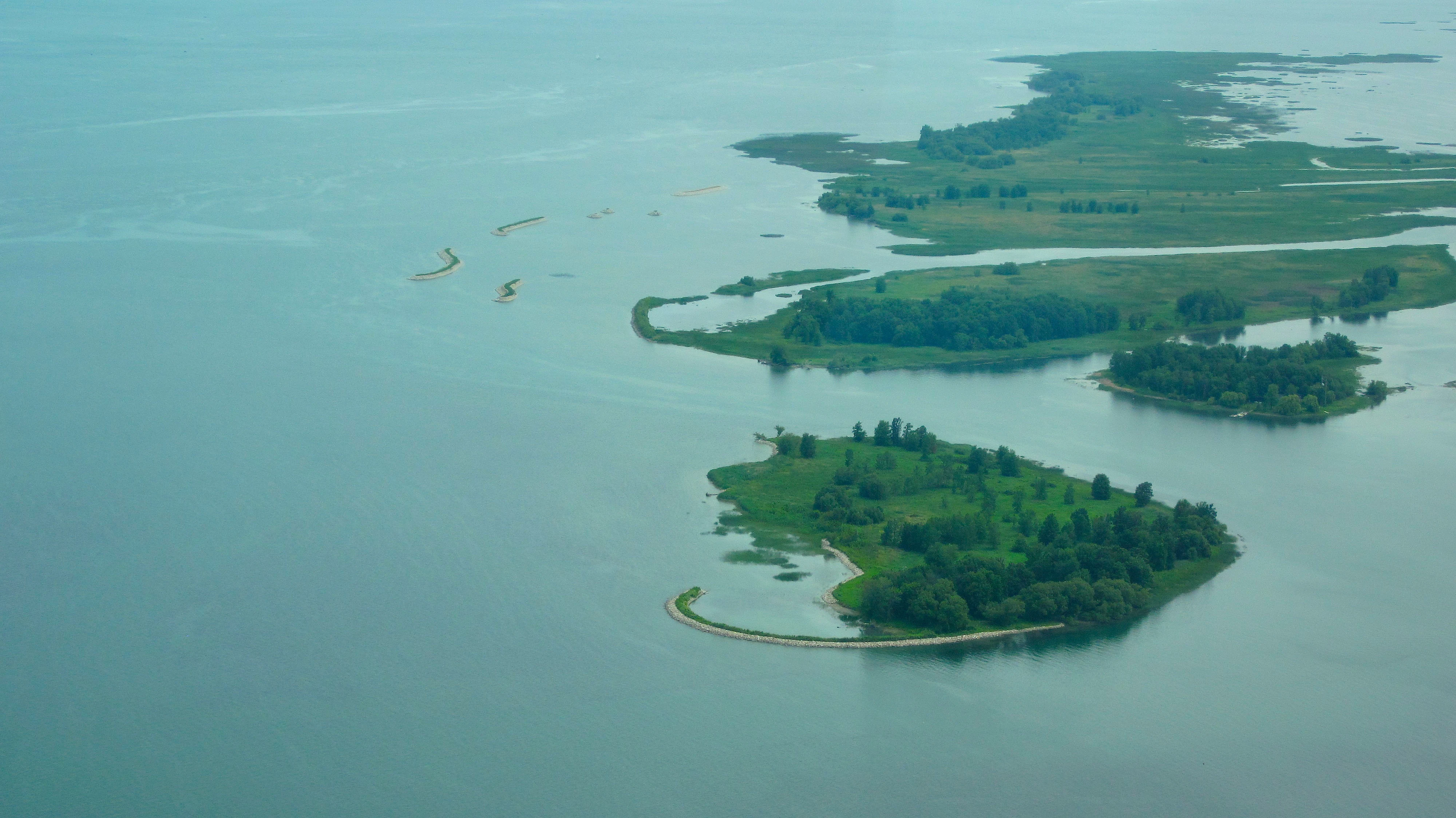 Aerial view of green islands surrounded by water, with visible shoreline protection structures like curved breakwaters off the coast of the islands.