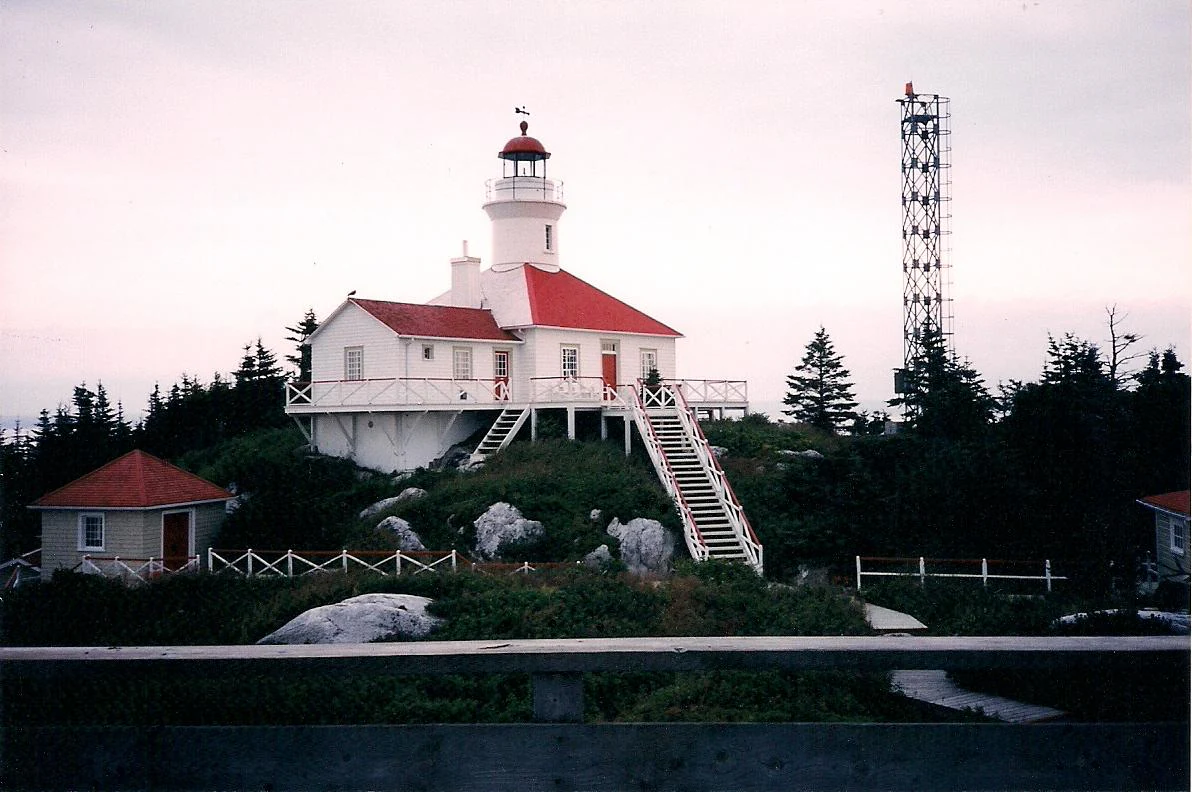 A white lighthouse with a red roof stands on a rocky island surrounded by evergreen trees, with a metal communications tower nearby.