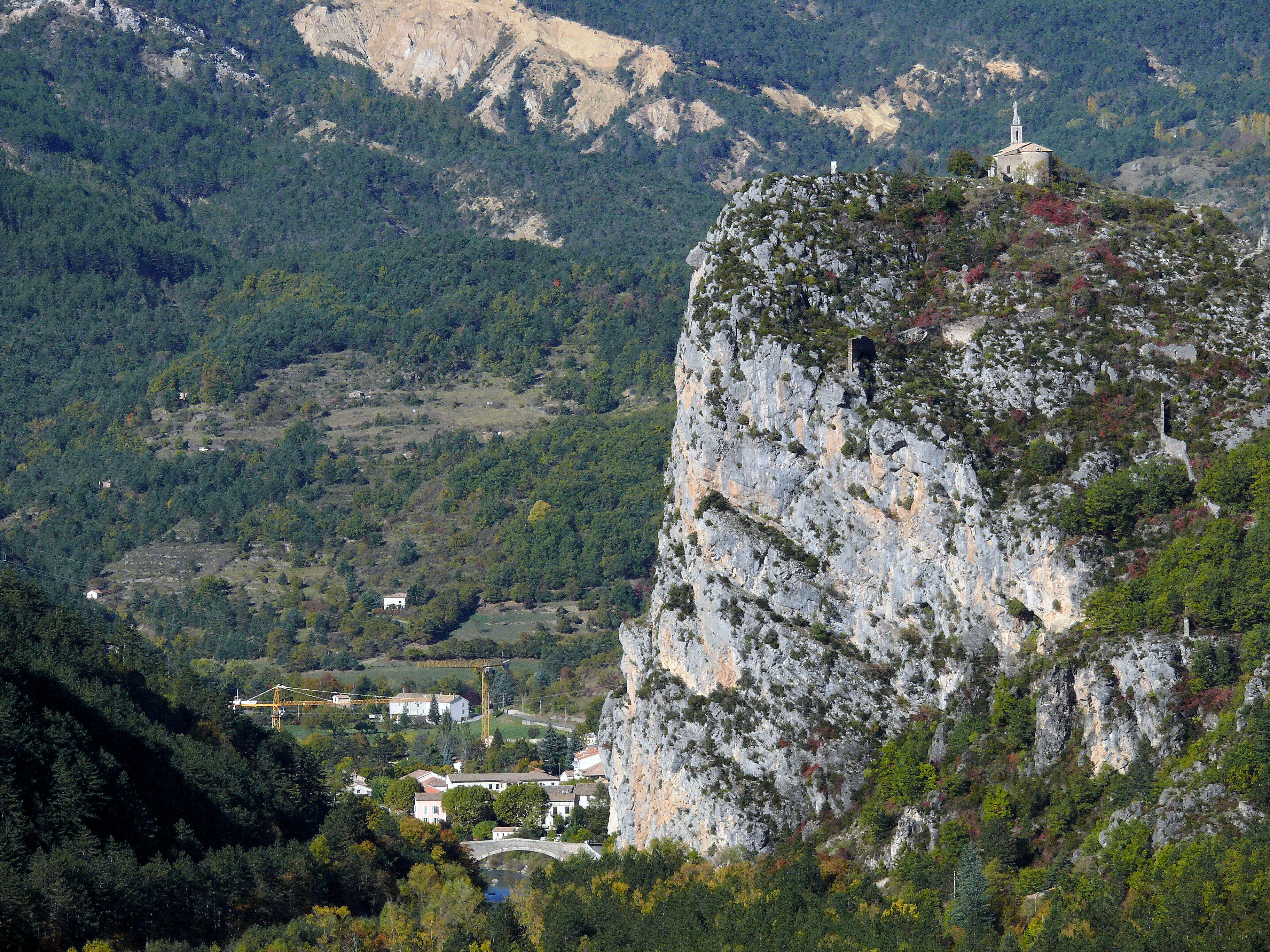 A large vertical limestone cliff, known as the Roc of Castellane, rises above a valley floor with dense green forest and a small cluster of buildings below.