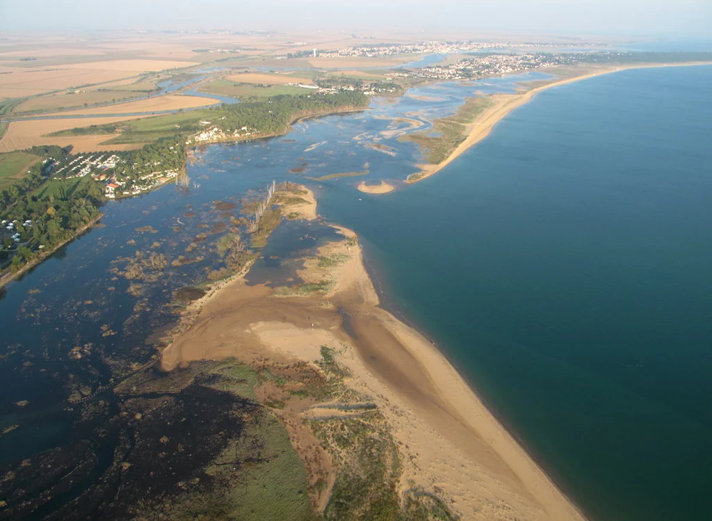 An aerial view of the Belle-Henriette lagoon showing a narrow sand spit separating the lagoon from the open sea, with marshes and surrounding coastal land.