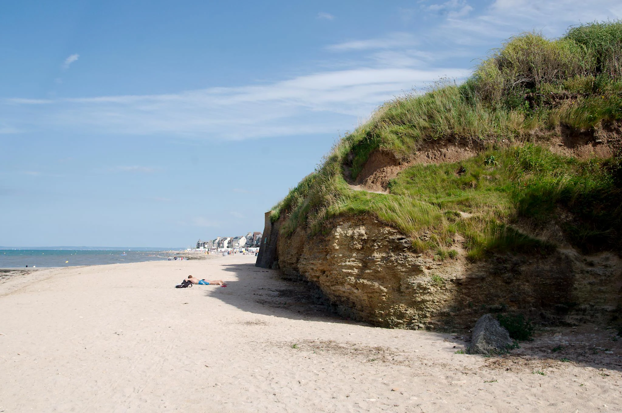 A wide sandy beach meeting a grassy, sedimentary cliff face under a blue sky.