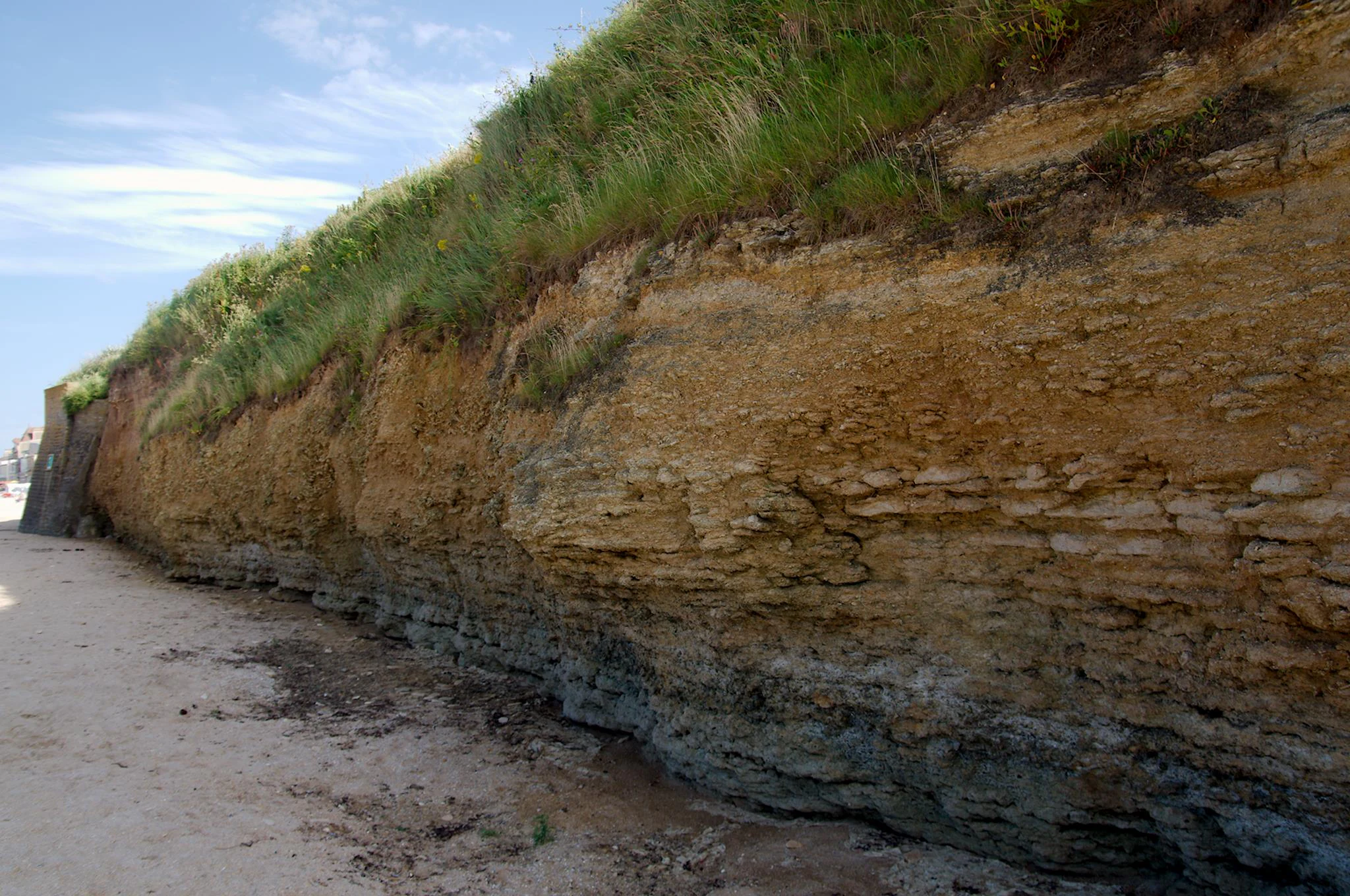 A low-angle view along the base of a coastal cliff showing distinct geological layers of yellow limestone and grey clay topped with green grass, with a sandy beach in the foreground.