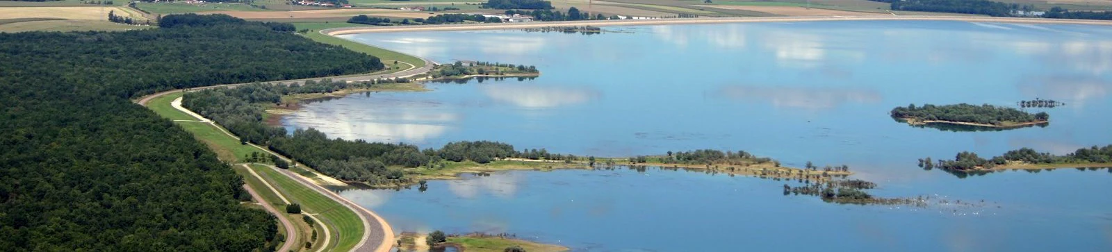 Aerial view of a large, blue lake bordered by a dense green forest and a man-made embankment with a path, under a clear sky.
