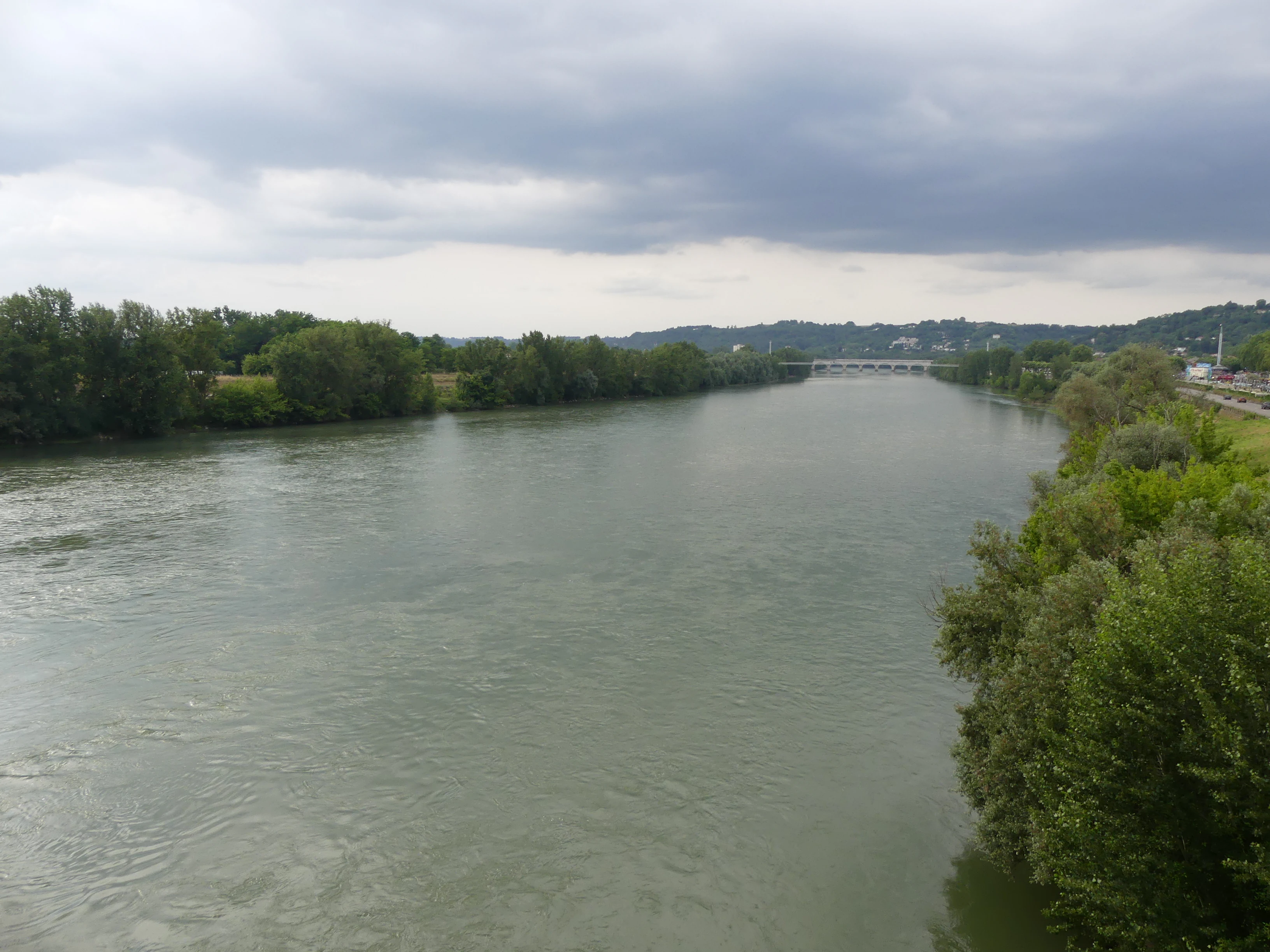 A wide, calm river flowing through a lush, tree-lined landscape under a cloudy sky.