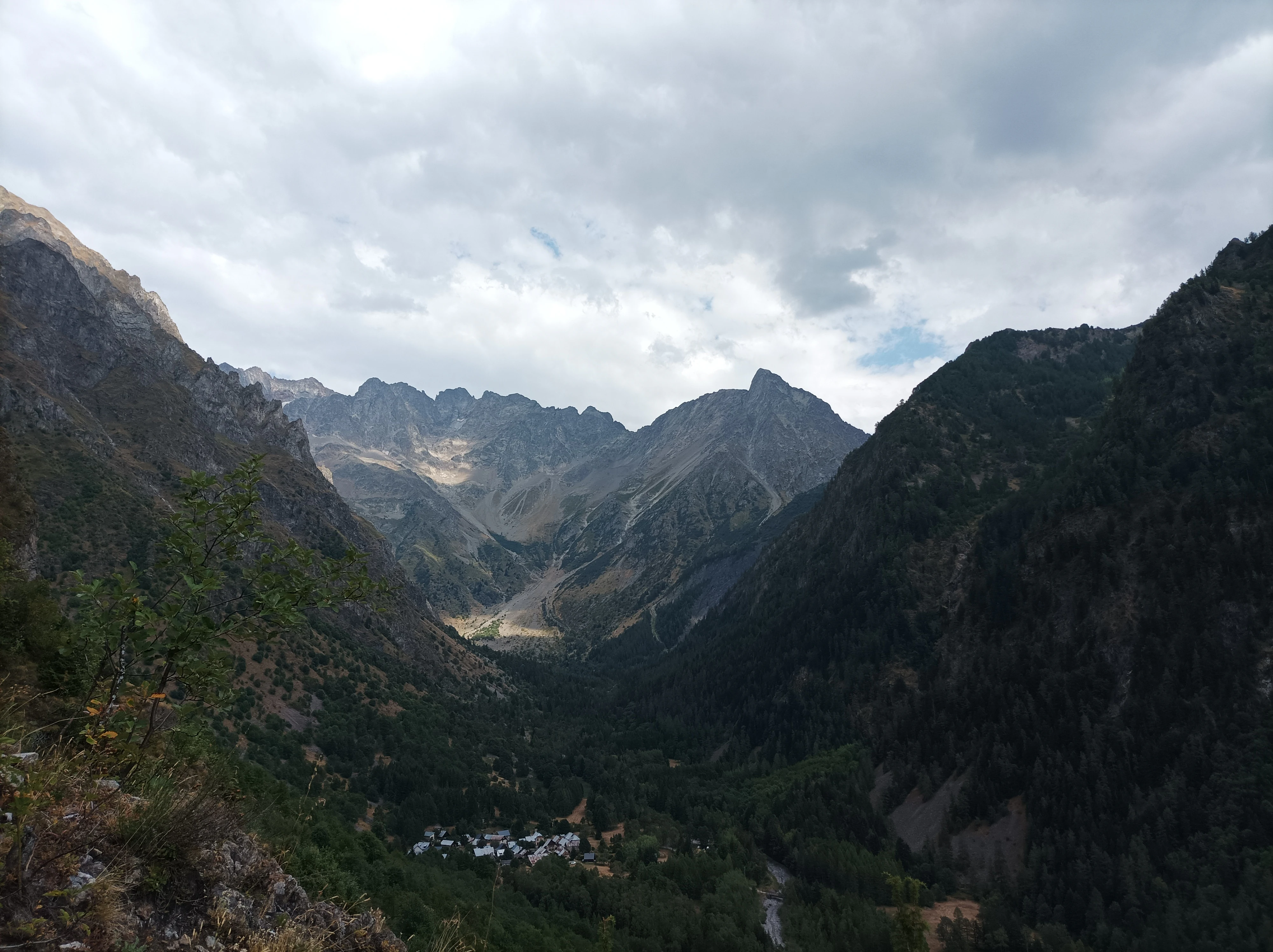 A wide, eye-level mountain landscape showing a steep, forested valley framed by high rocky peaks under a cloudy sky.