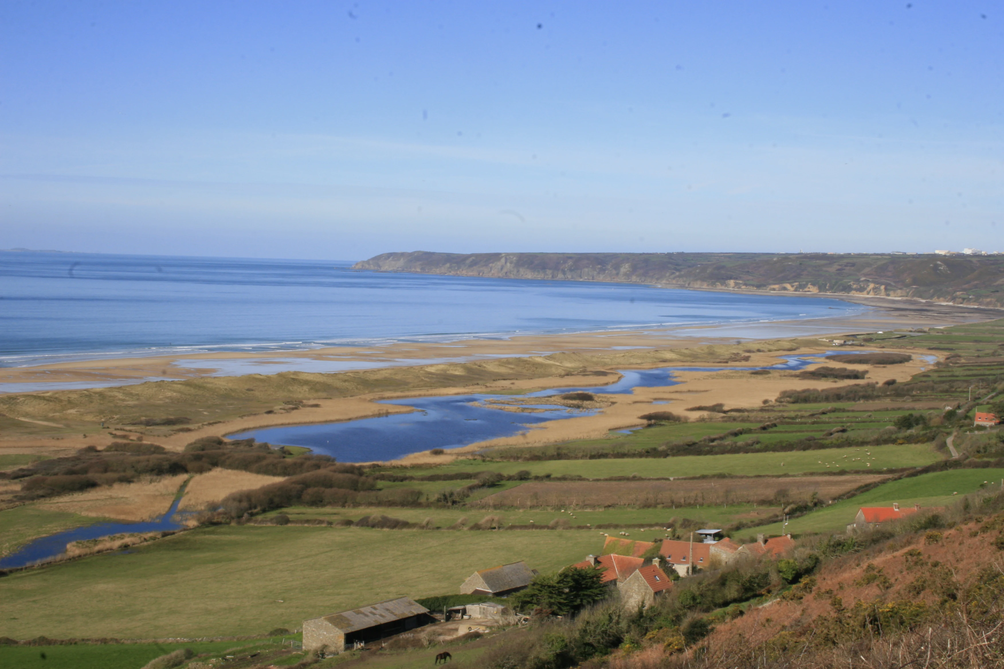 Una vista de gran angular de una reserva natural costera con una zona de marisma en primer plano, dunas de arena dorada en el plano medio y el mar encontrándose con el horizonte al fondo bajo un cielo despejado.