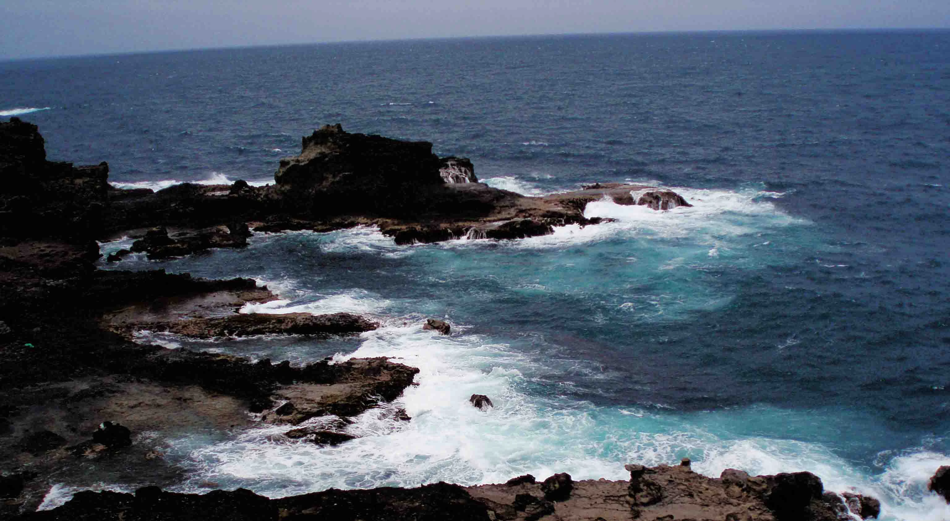 A high-angle view of rugged dark volcanic rocks along a coastline with white sea foam breaking against the shore in the Atlantic Ocean.