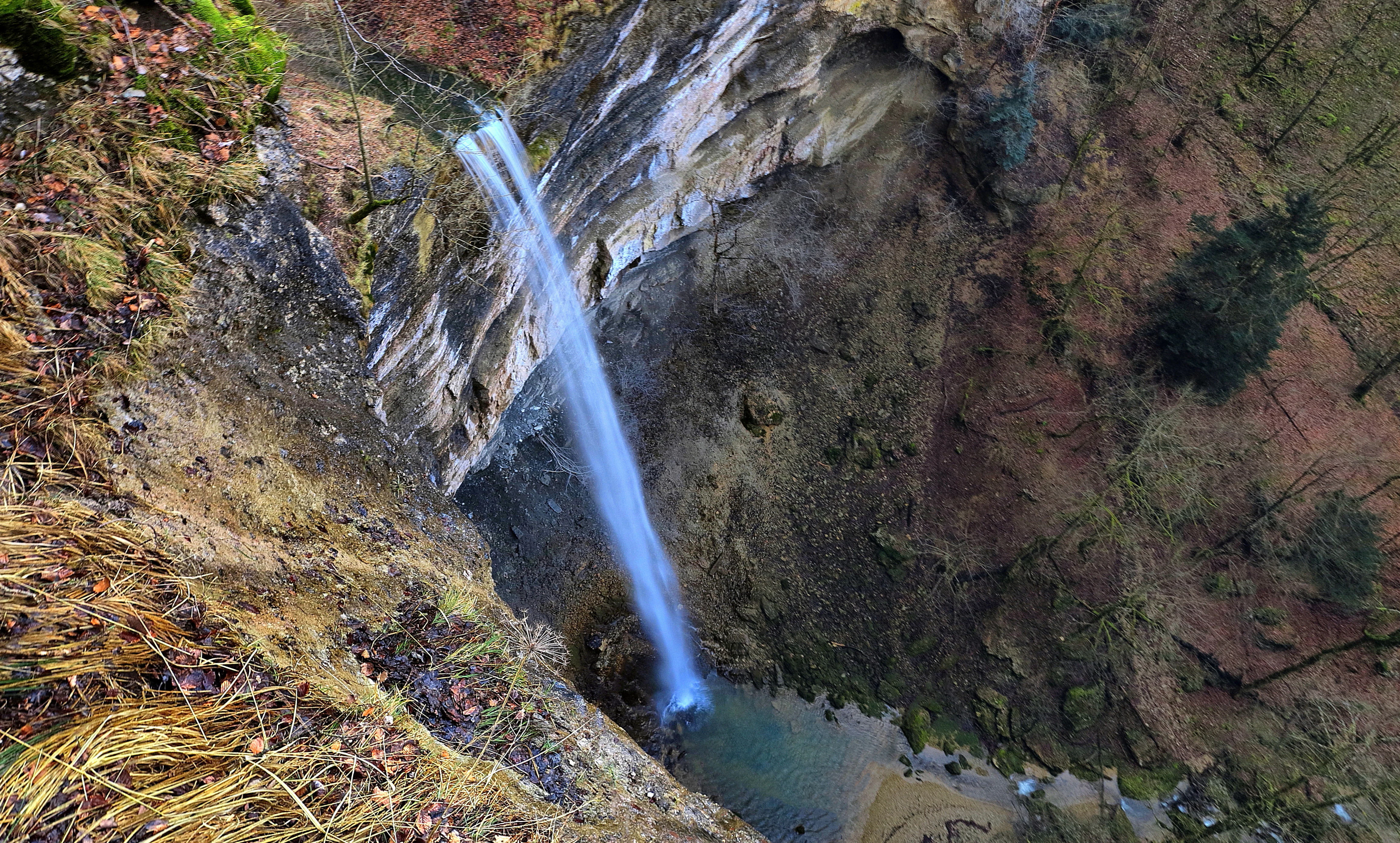 A high angle view looking down at a slender waterfall plunging over a rocky, limestone cliff face into a small pool, surrounded by forest vegetation.