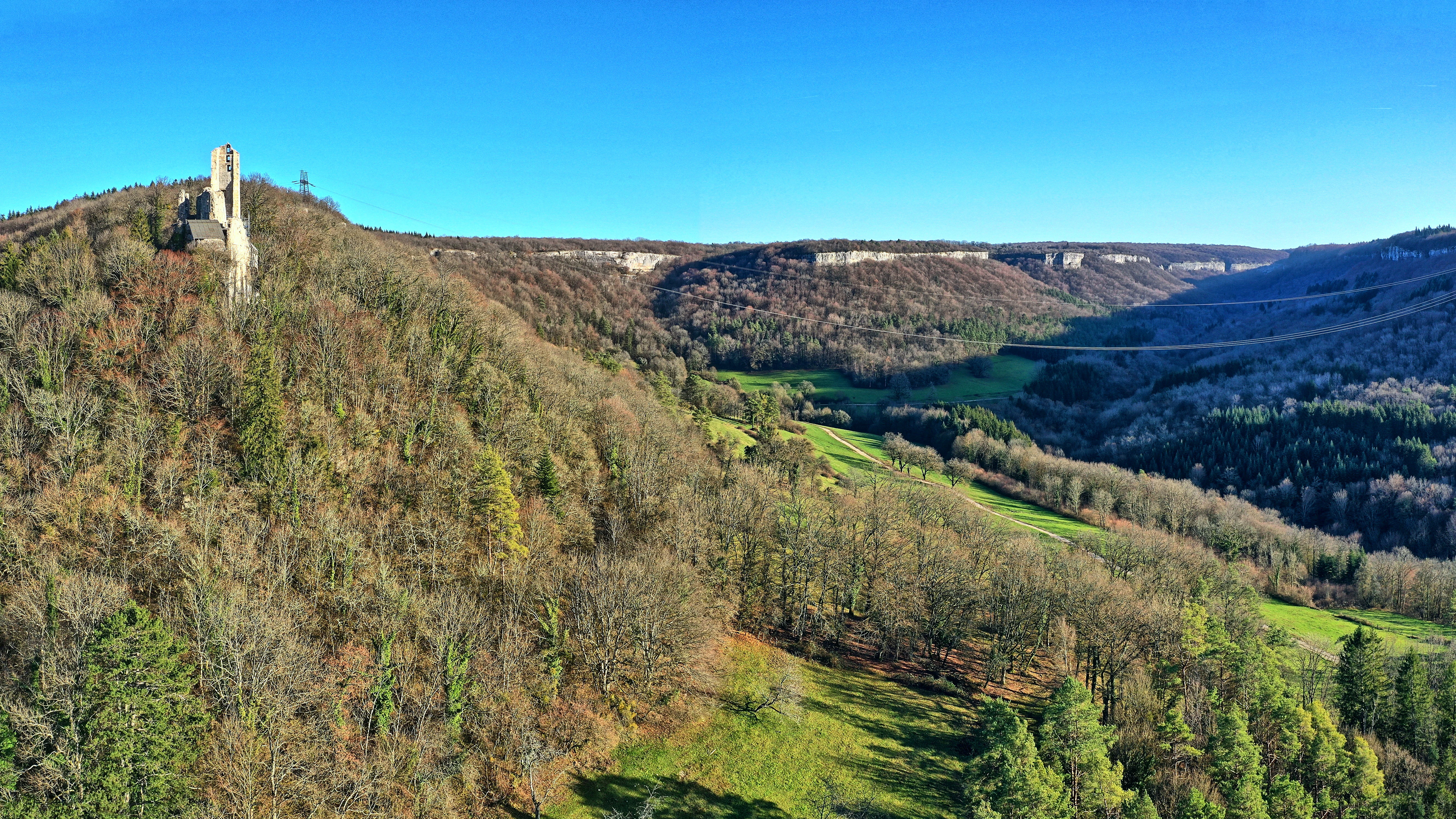 An elevated view of a forested ravine landscape with a ruined stone castle tower perched on a hillside in the foreground and limestone cliffs lining the valley.
