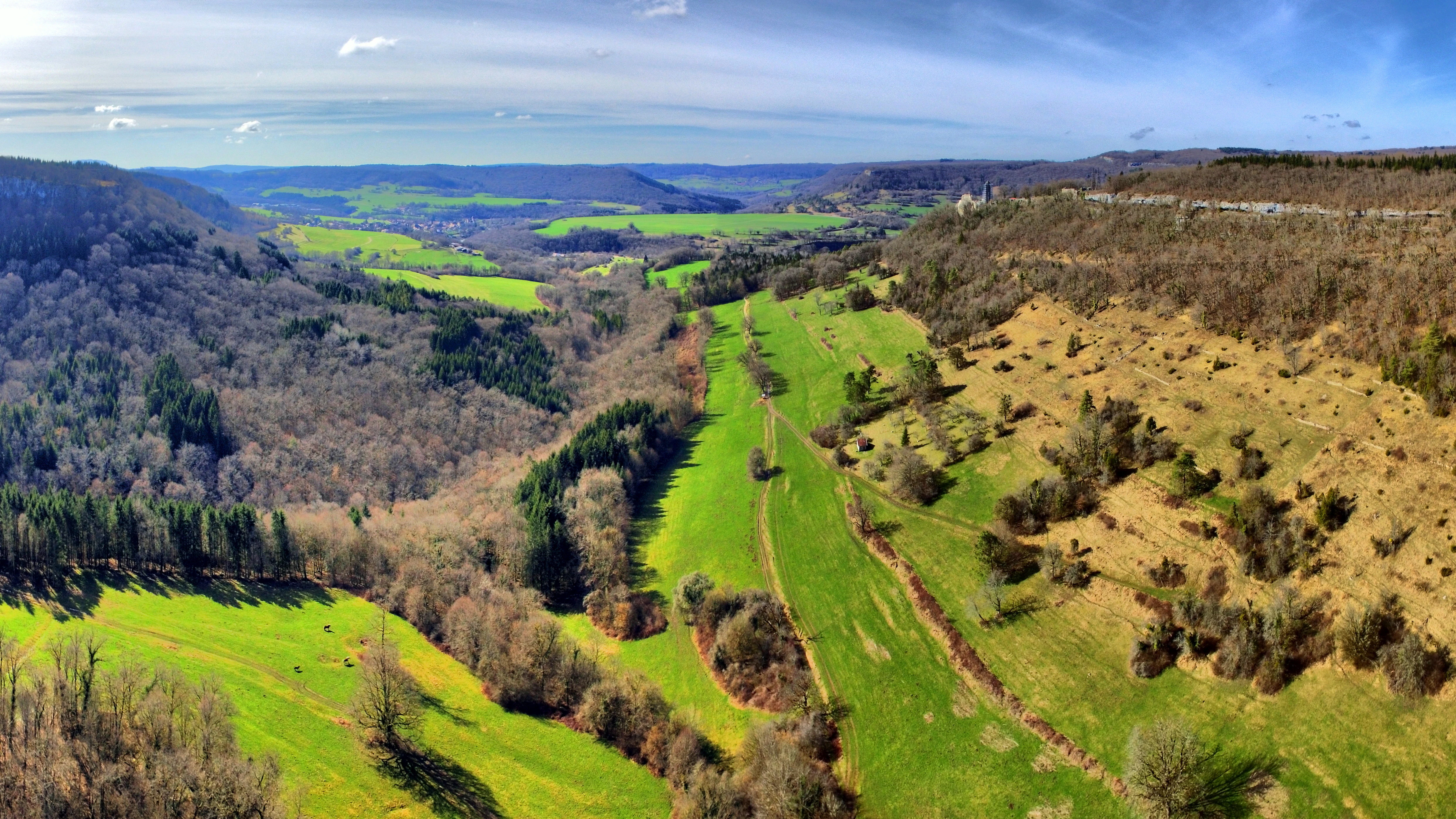 A high-angle panoramic landscape view showing a green valley merging with a larger valley, characterized by mixed forests and limestone slopes