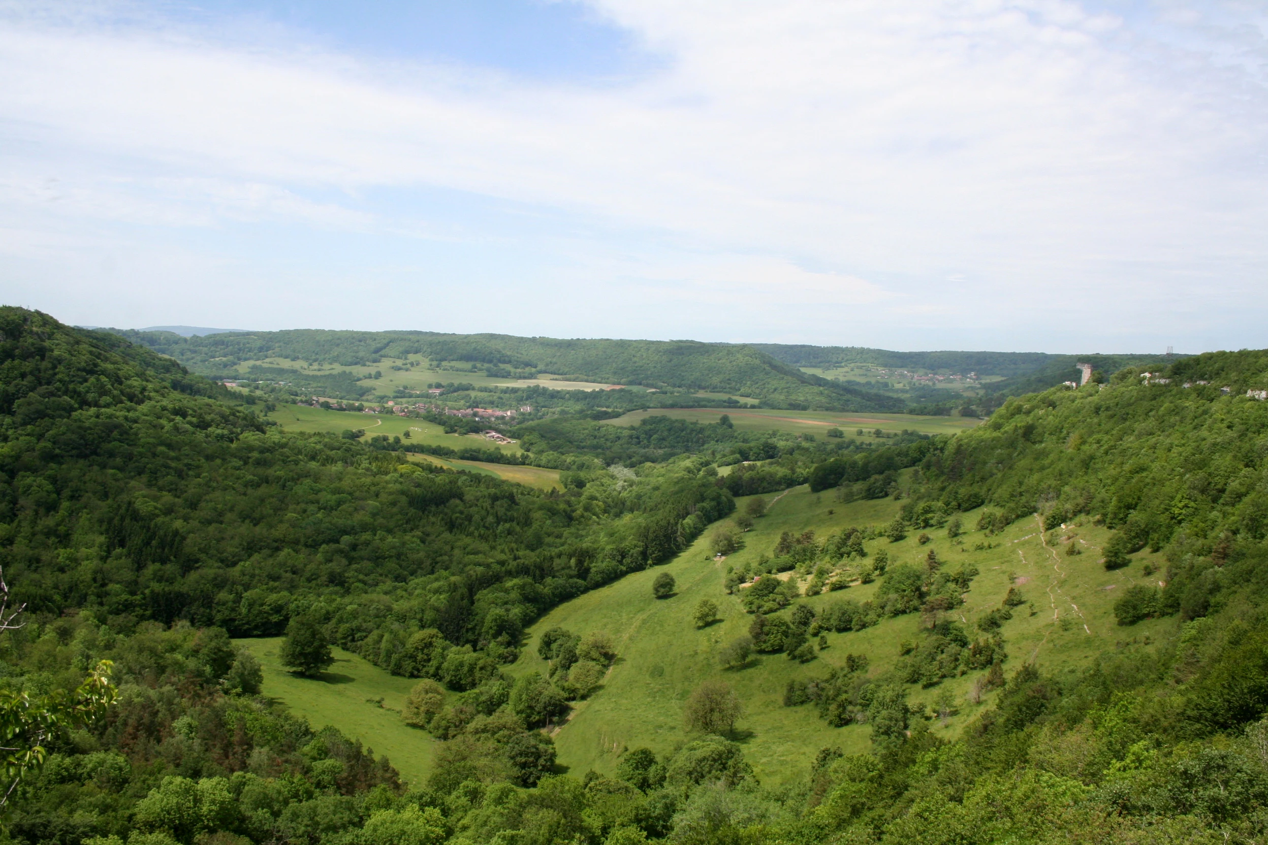 A wide, high-angle view overlooking a lush, green, U-shaped valley with steep, forested cliffs on the sides and a gentle grassy slope in the foreground under a light blue sky with wispy clouds.