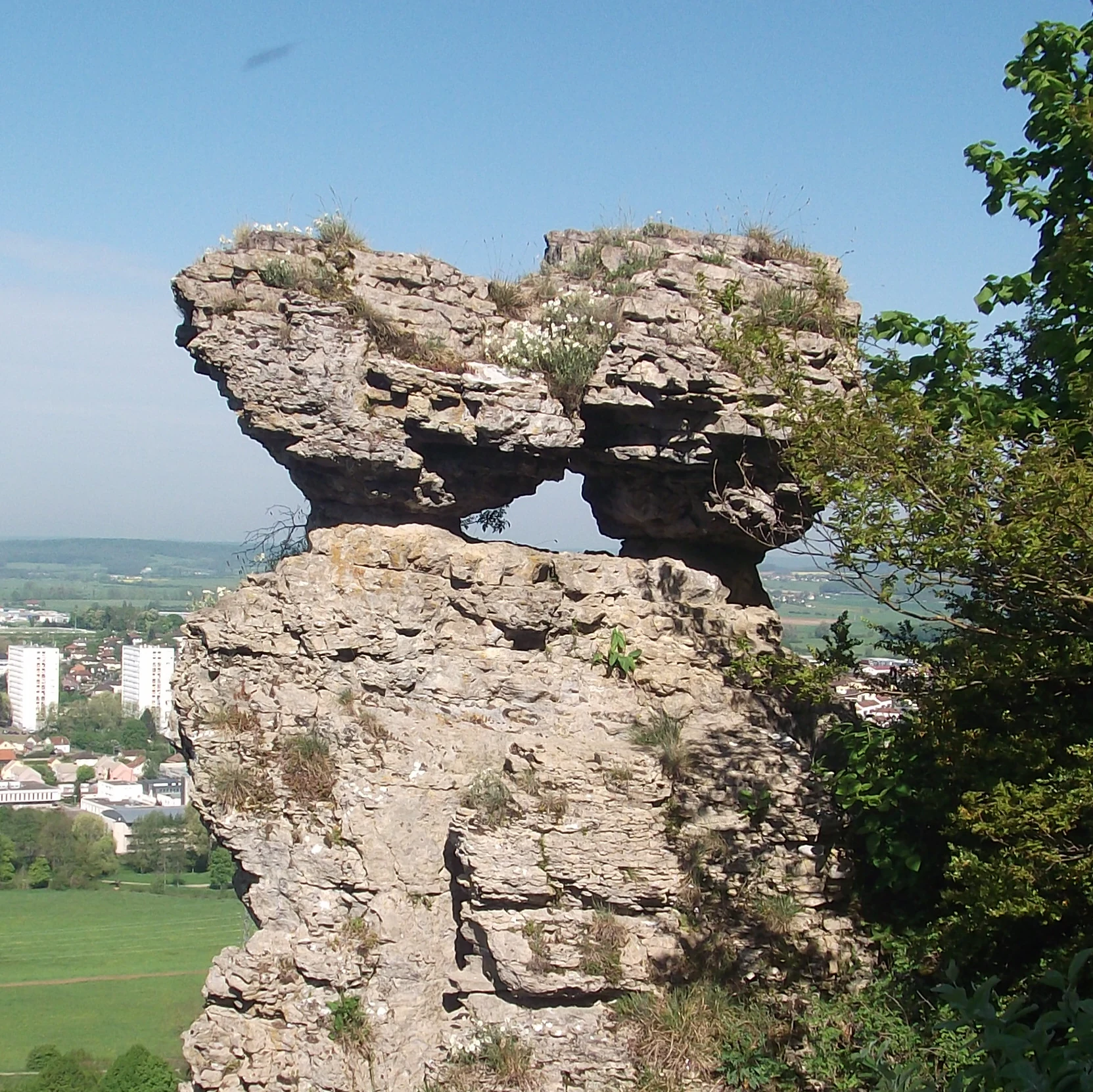A large, weathered limestone rock formation shaped like a clog or mushroom, with a small hole in its center, overlooking a town and green valley in the distance.