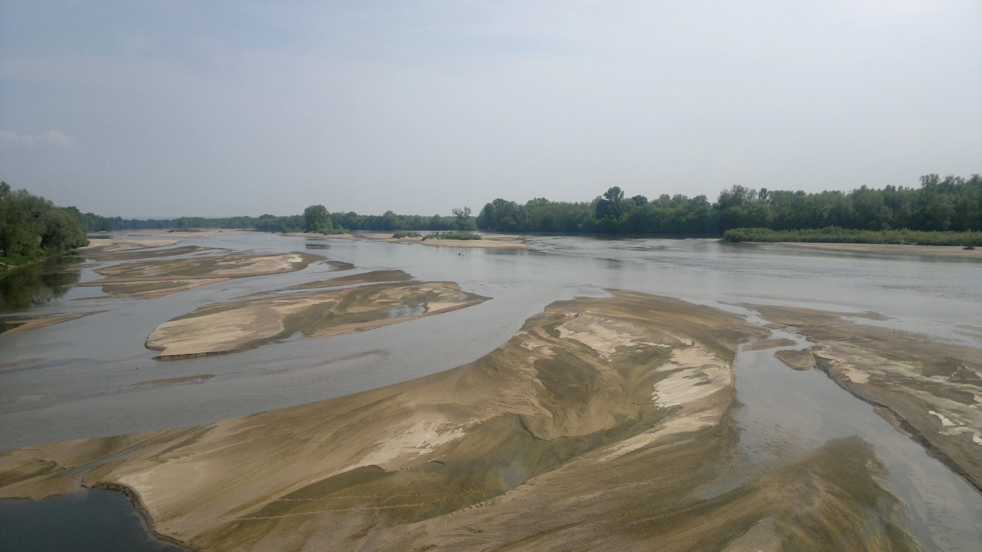 Wide angle view of the Loire River with numerous exposed sandbars and shallow water channels, framed by green trees on the banks under a hazy sky.