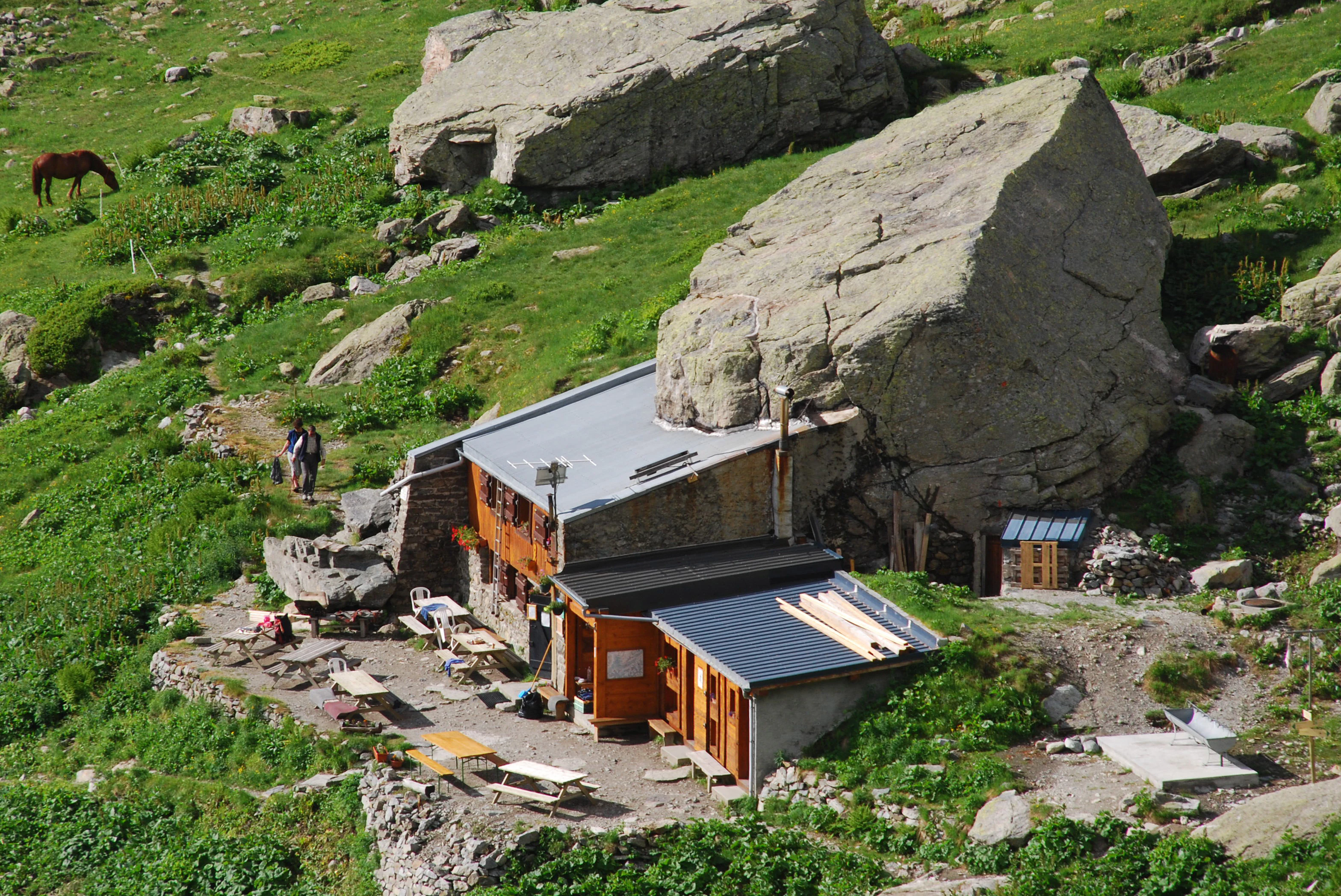 High angle shot of a wooden and stone mountain refuge built directly beneath a massive boulder in a lush green alpine meadow, with a horse grazing nearby.