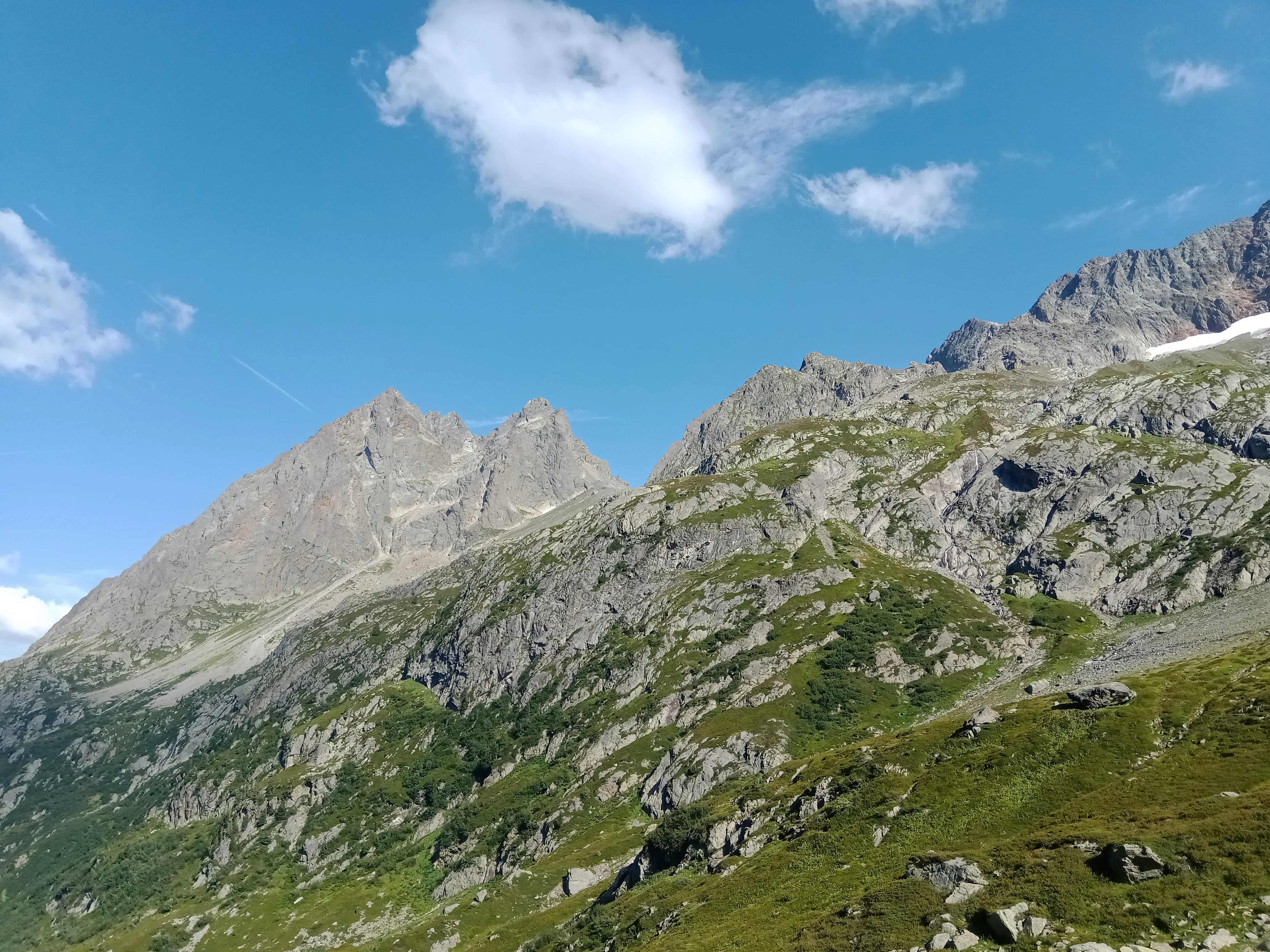 A wide shot of steep, green, and rocky alpine mountain slopes under a clear blue sky with light clouds.