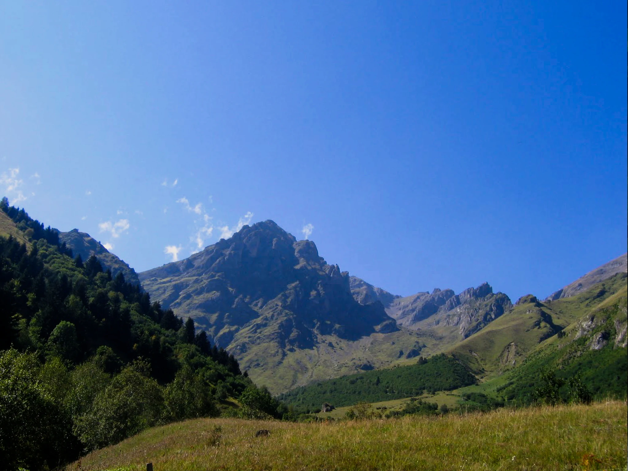 A clear, sunny day view of a rugged, rocky mountain peak, the Mailh d'Aulon, rising above a green valley and forest slopes.