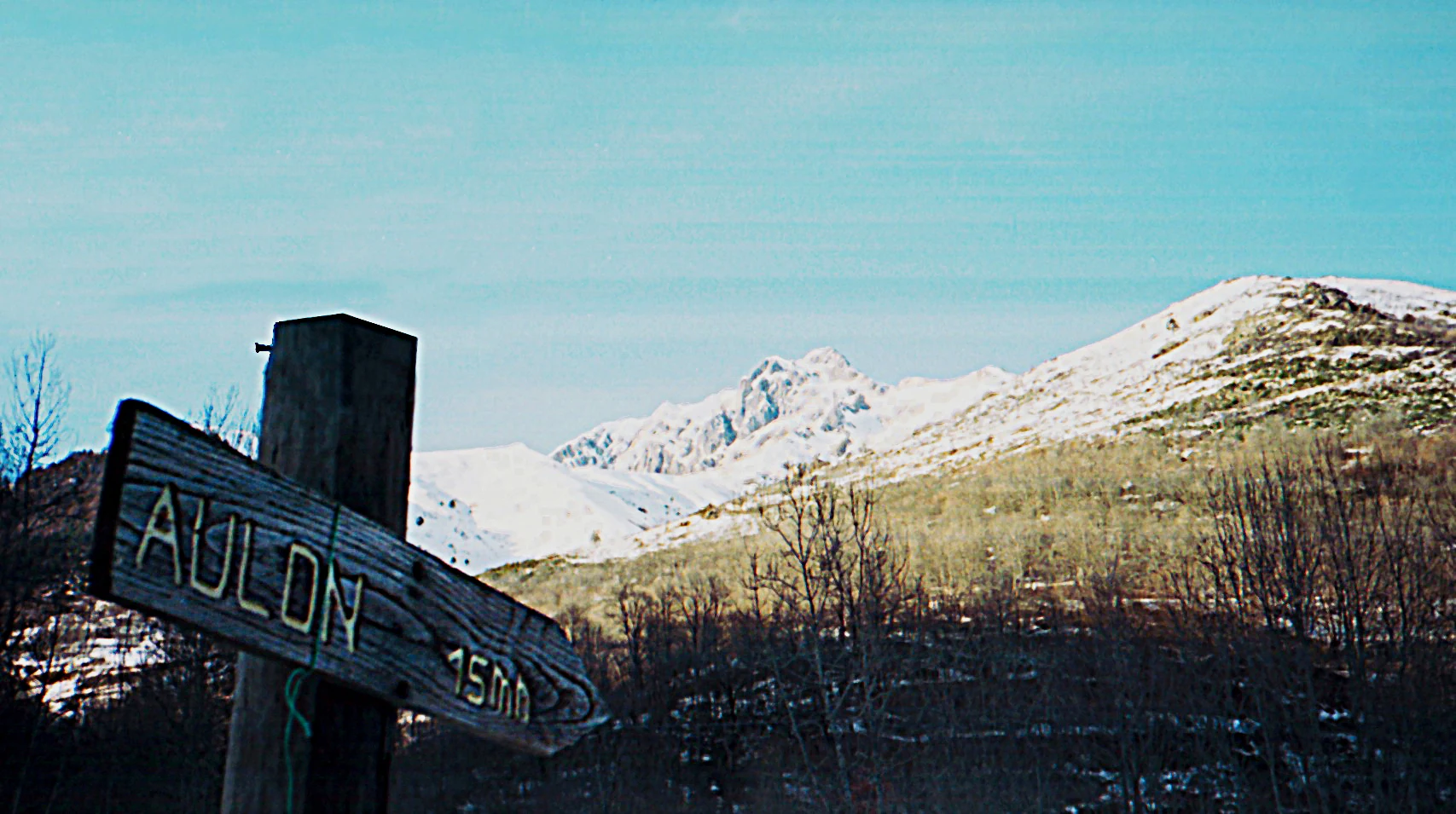 A weathered wooden hiking signpost with the text Aulon 15 min pointing to the left, set against a backdrop of snow-covered Pyrenean mountains and sparse winter forest.