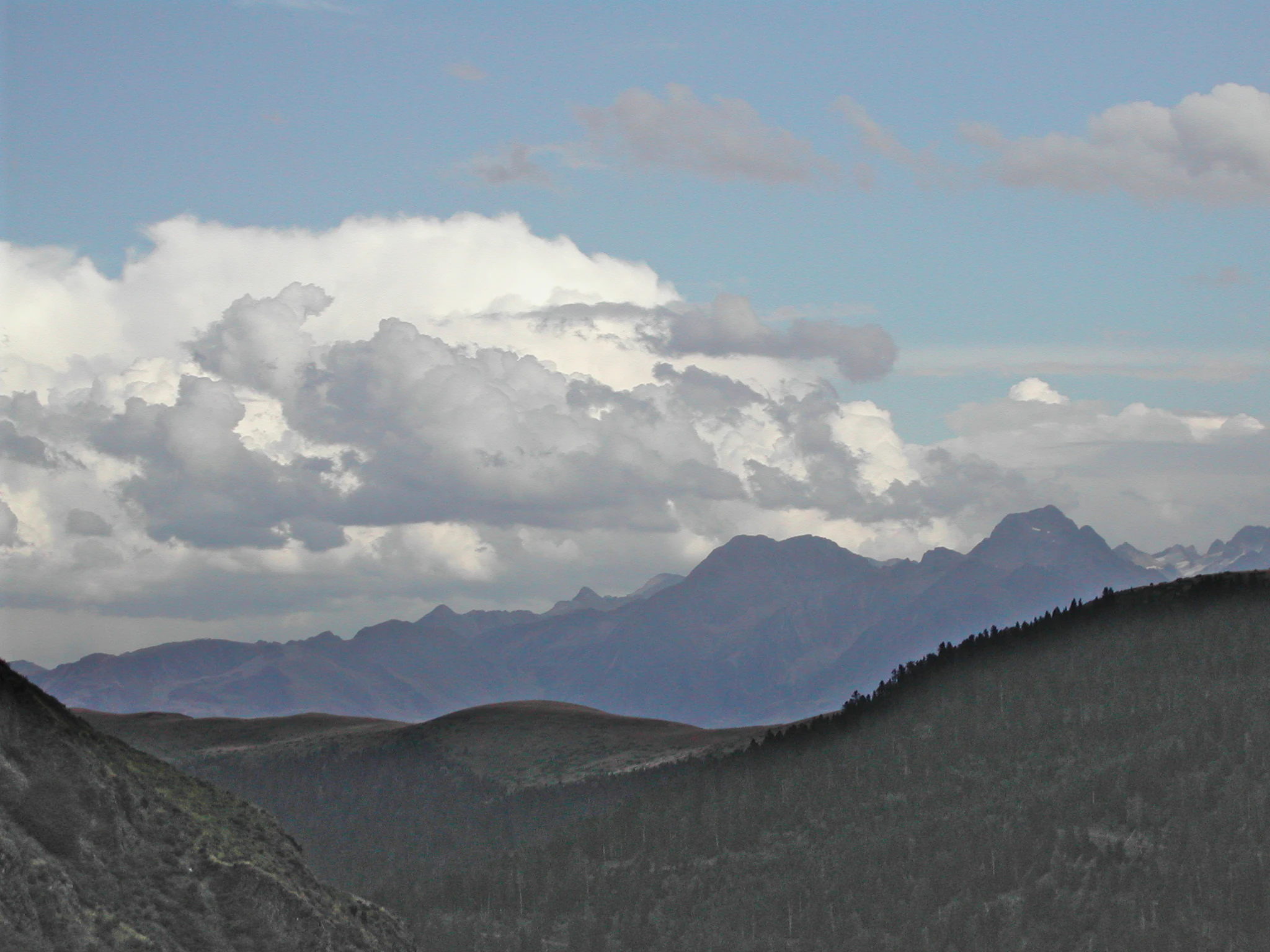 A wide mountain landscape showing a series of distant rugged peaks under a partly cloudy sky, with dark, shadowed mountain slopes framing the view in the foreground.