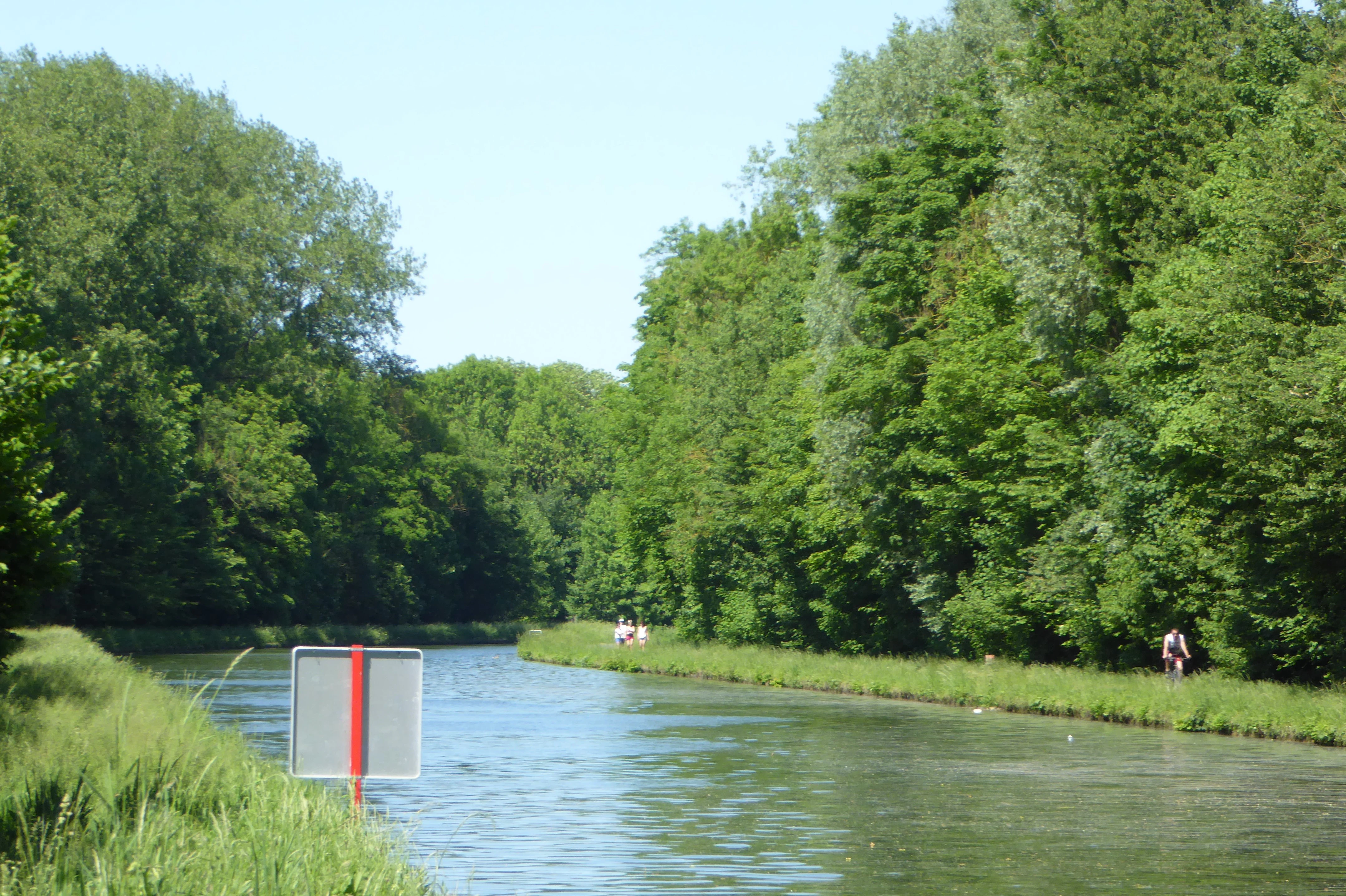 A peaceful canal or river flowing through a lush green forest, with a metal sign visible in the foreground and a grassy towpath on the right.