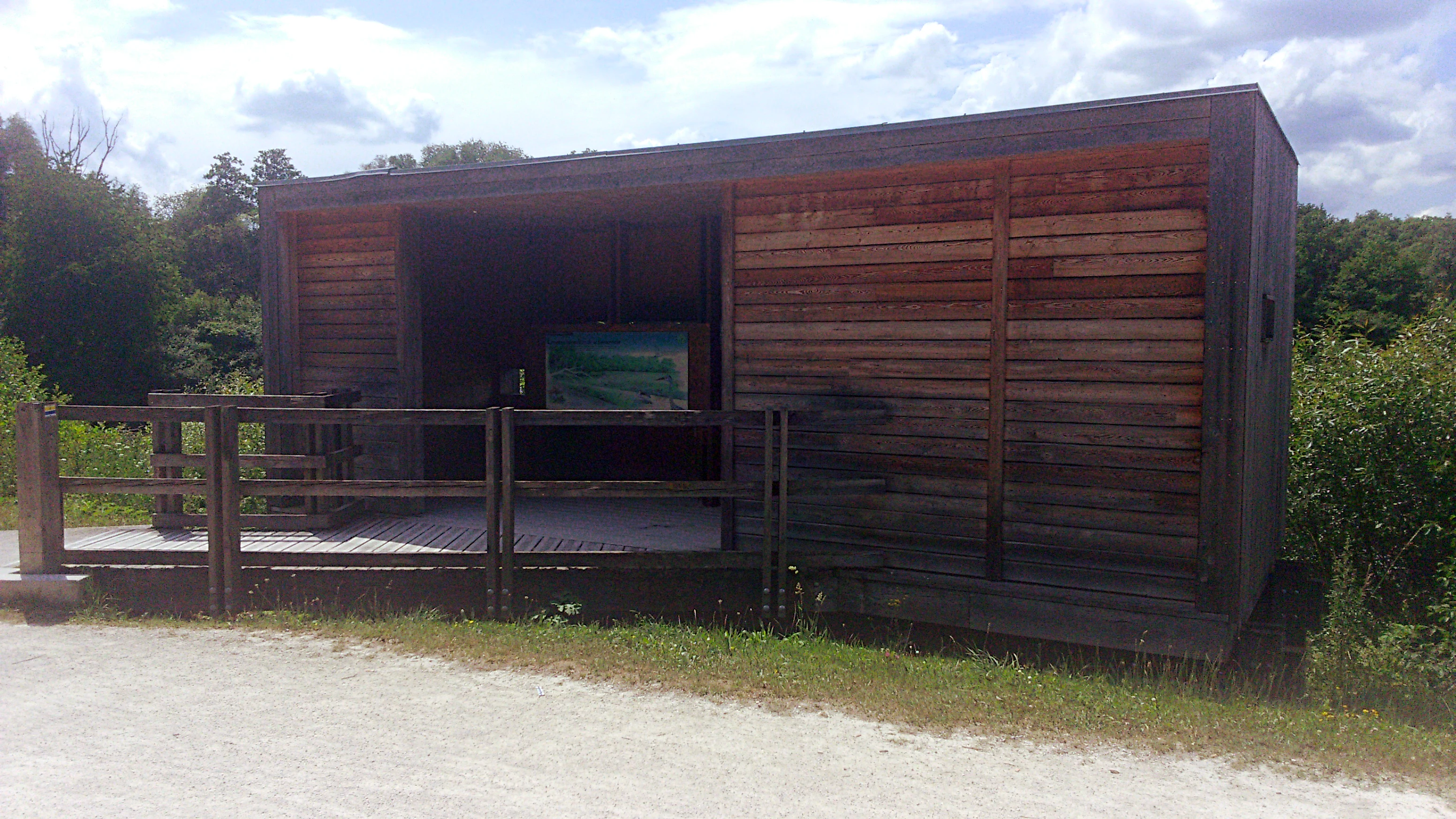 A wooden observation structure with horizontal slat cladding, featuring an open viewing area with a wooden deck and railing, surrounded by green vegetation under a partly cloudy sky