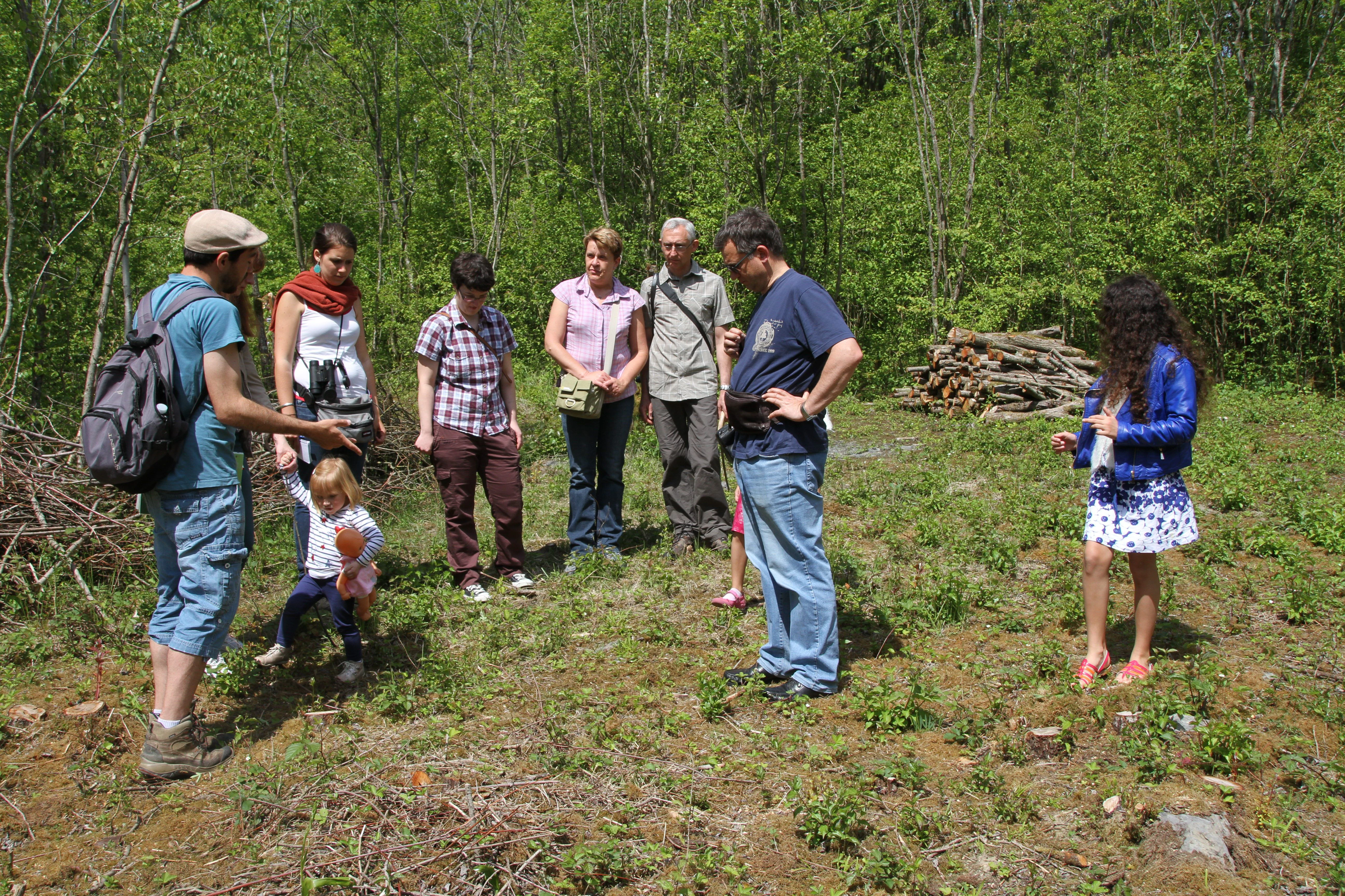 A group of adults and a child gathered in a grassy clearing with a wooded background, listening to a guide during an outdoor nature tour.