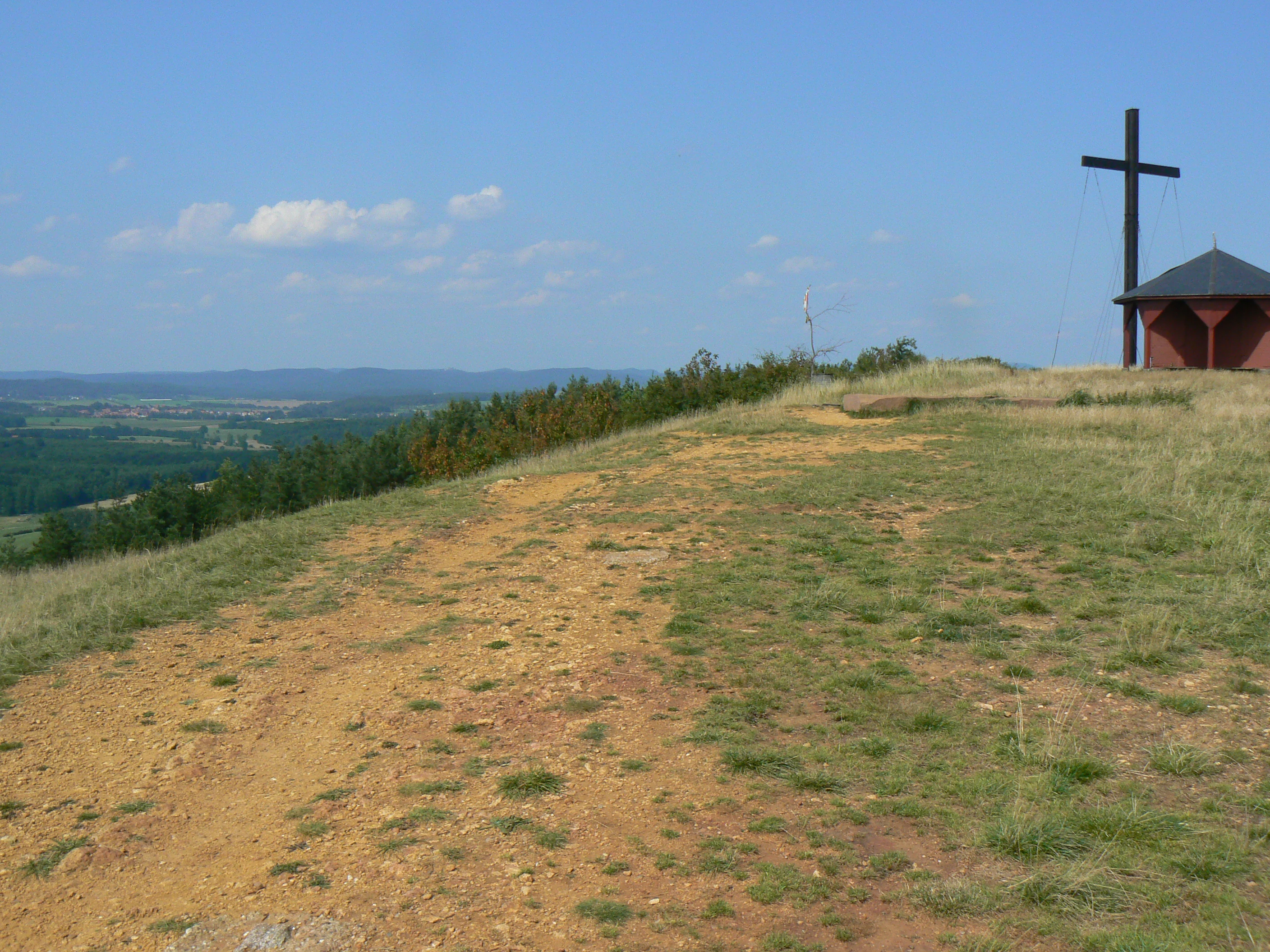 A grassy hilltop with a tall dark cross and a small wooden building on the right, overlooking a vast landscape under a clear blue sky.