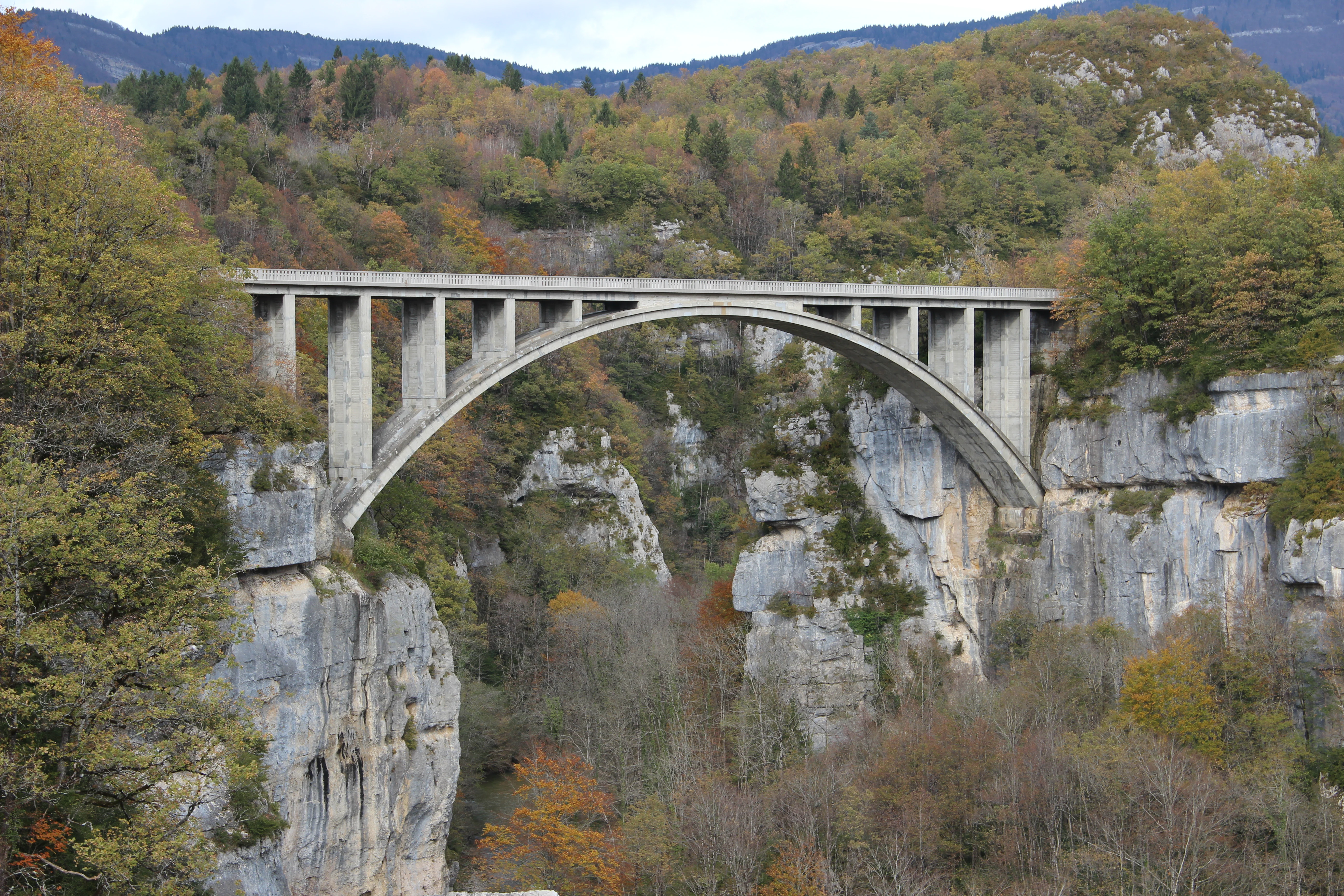 Un gran puente de arco de hormigón cruza un profundo desfiladero rocoso rodeado de follaje otoñal en un valle boscoso.