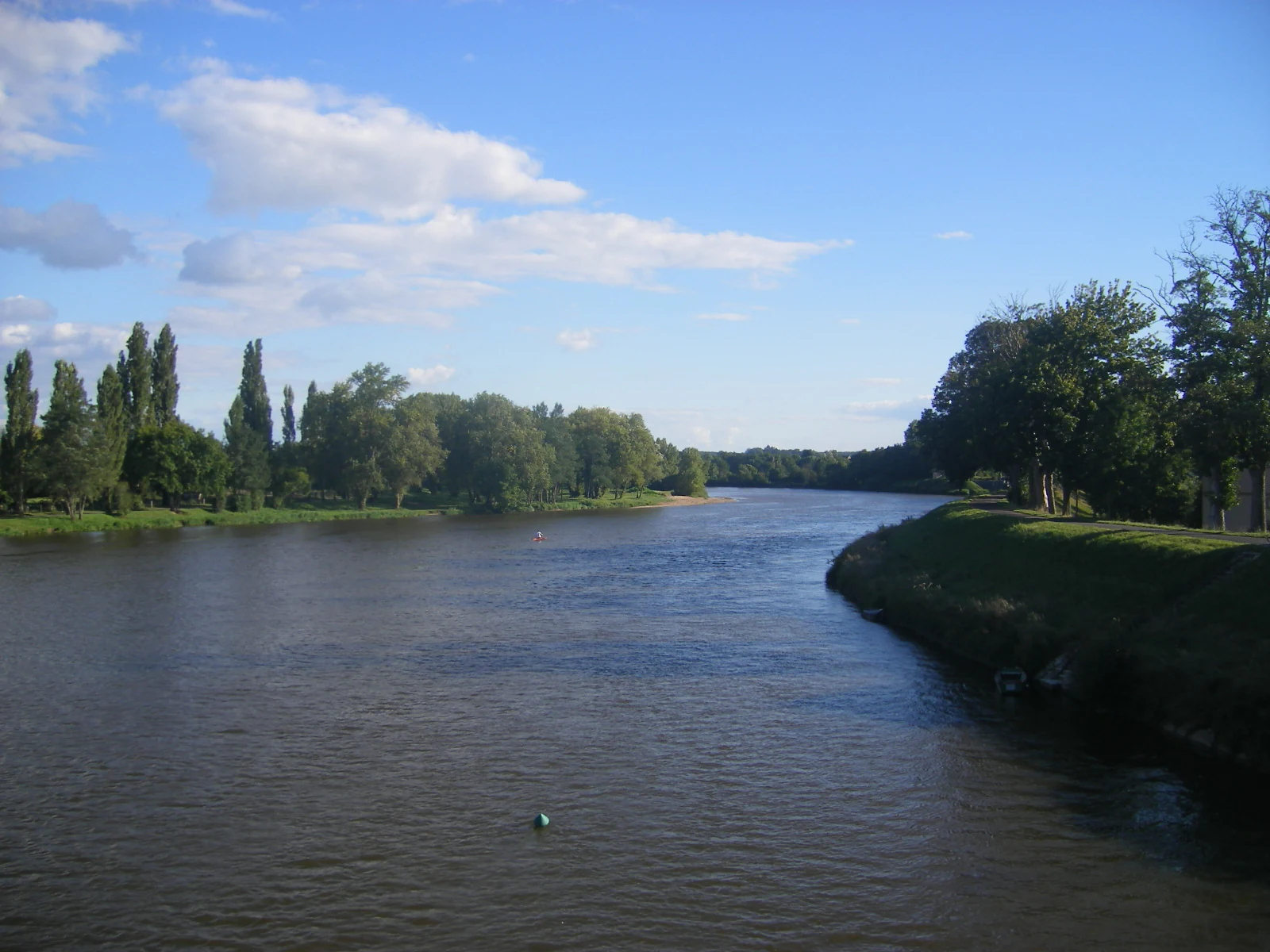 A wide view of a calm, wide river flowing through a lush green valley with trees lining both banks under a bright blue sky.
