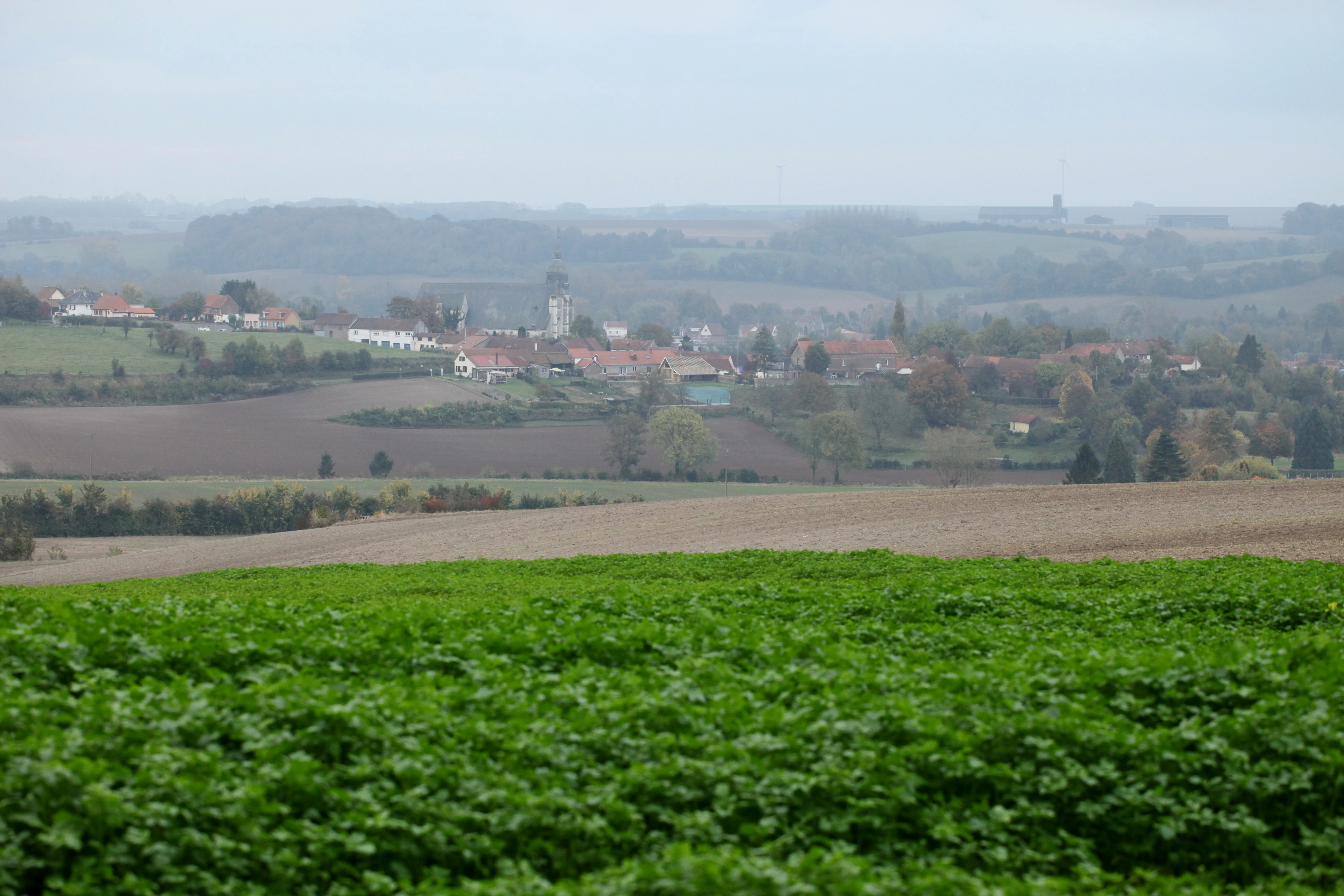 Una vista amplia y brumosa de un pequeño pueblo francés enclavado entre campos ondulados y colinas, tomada desde un punto elevado en la reserva de Pâture Mille Trous.
