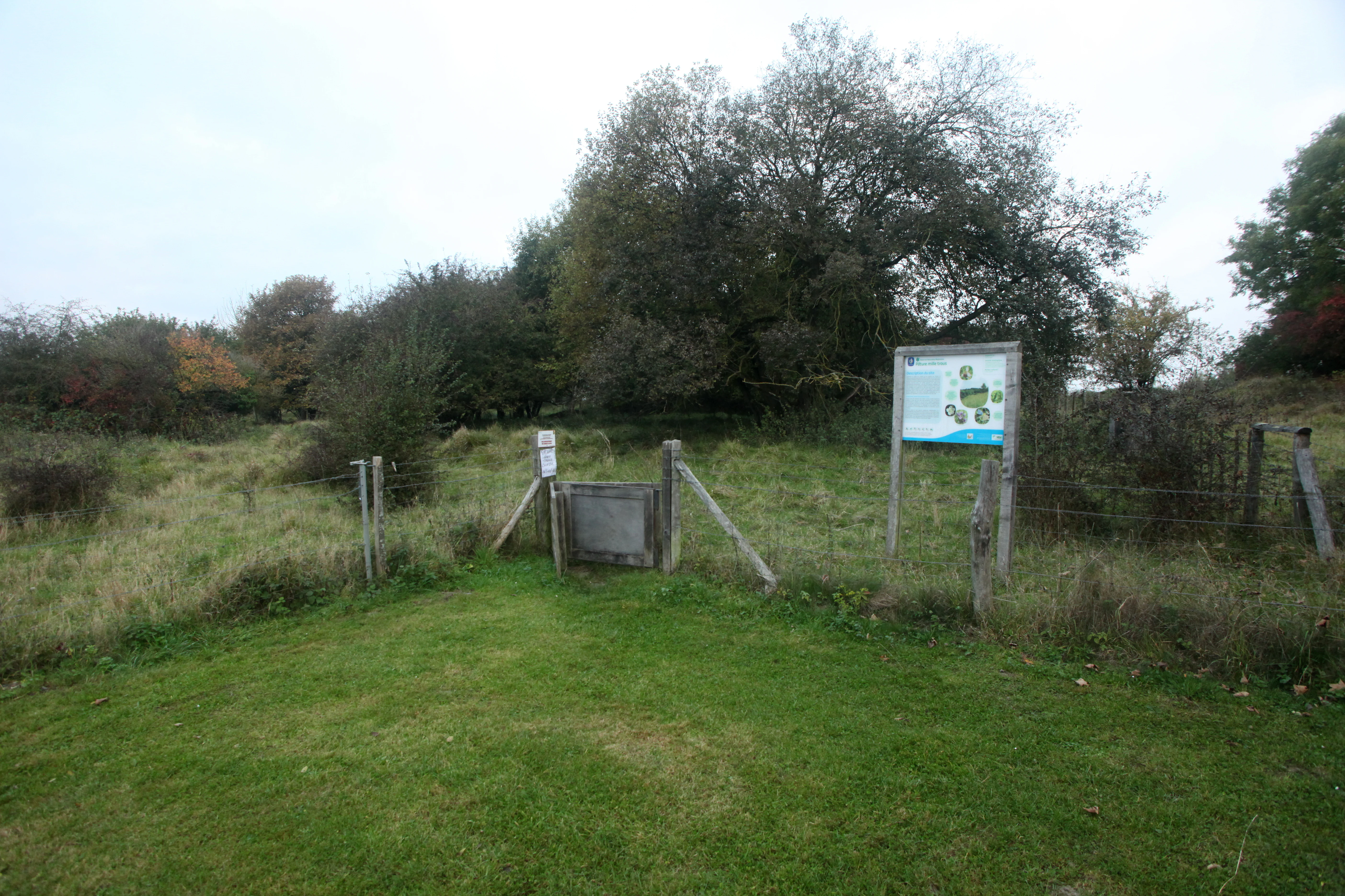 Una puerta de madera y un cartel informativo del parque marcan el punto de entrada a una reserva natural de pastizales con árboles al fondo.