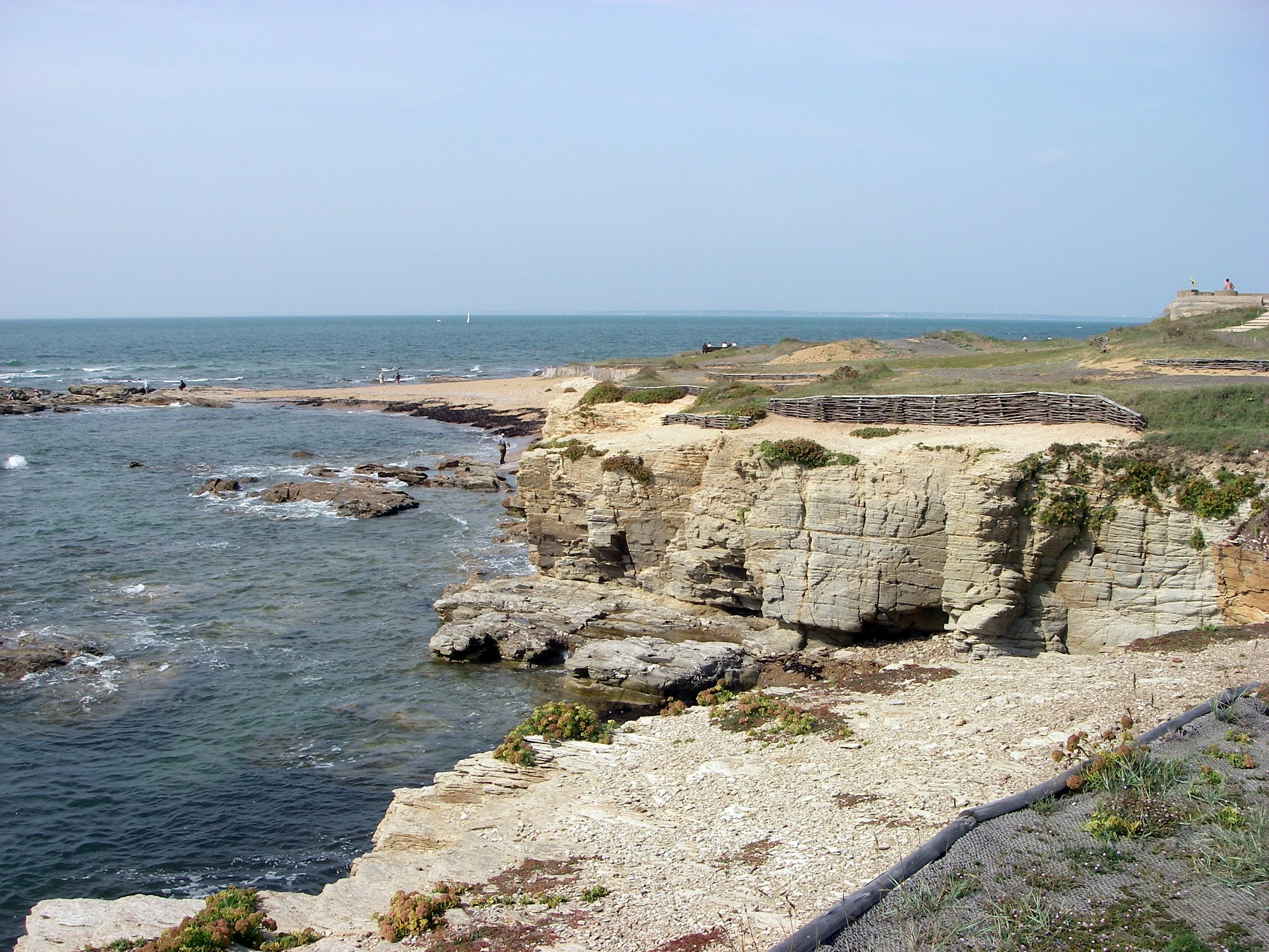 A wide view of a rocky coastline with light-colored cliffs meeting the sea, featuring a low wooden fence structure on the headland.