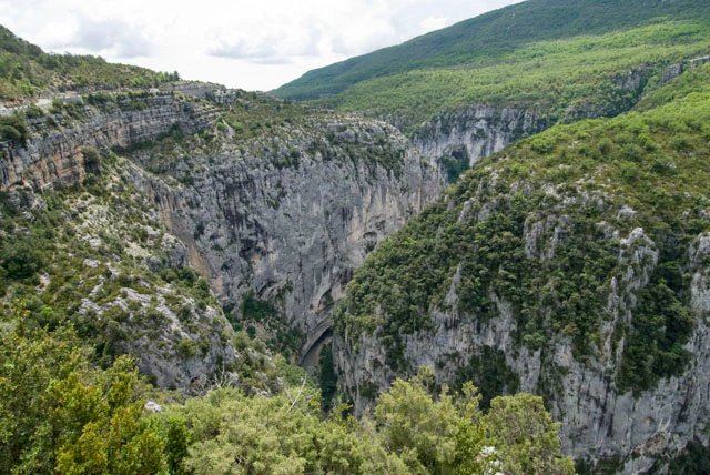 A wide, high-angle view looking down into a deep river gorge with sheer limestone cliffs covered in green Mediterranean scrub vegetation.