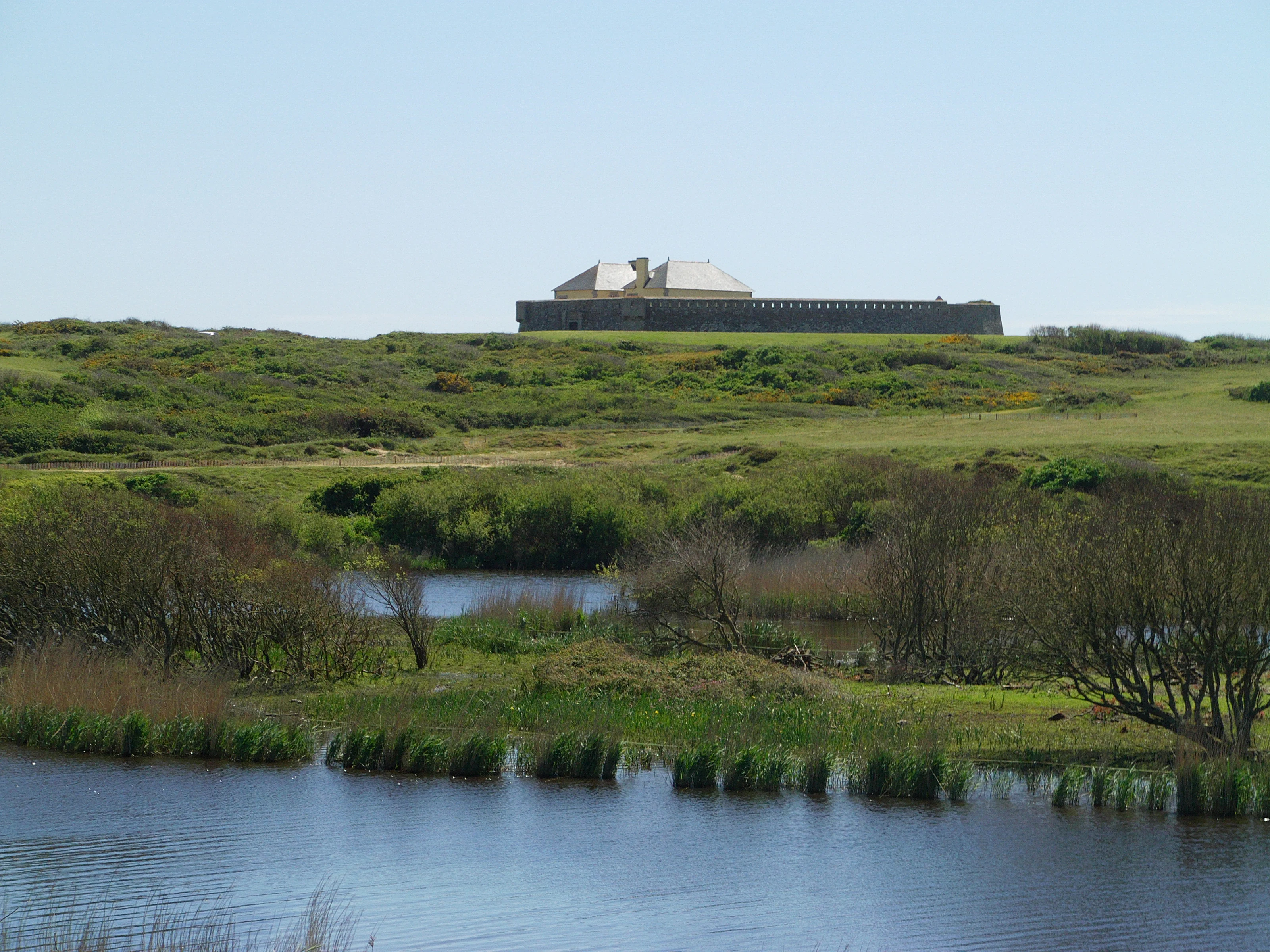 A wide view of a stone fort with a gabled roof sitting on a green grassy hill above a marshy pond area.