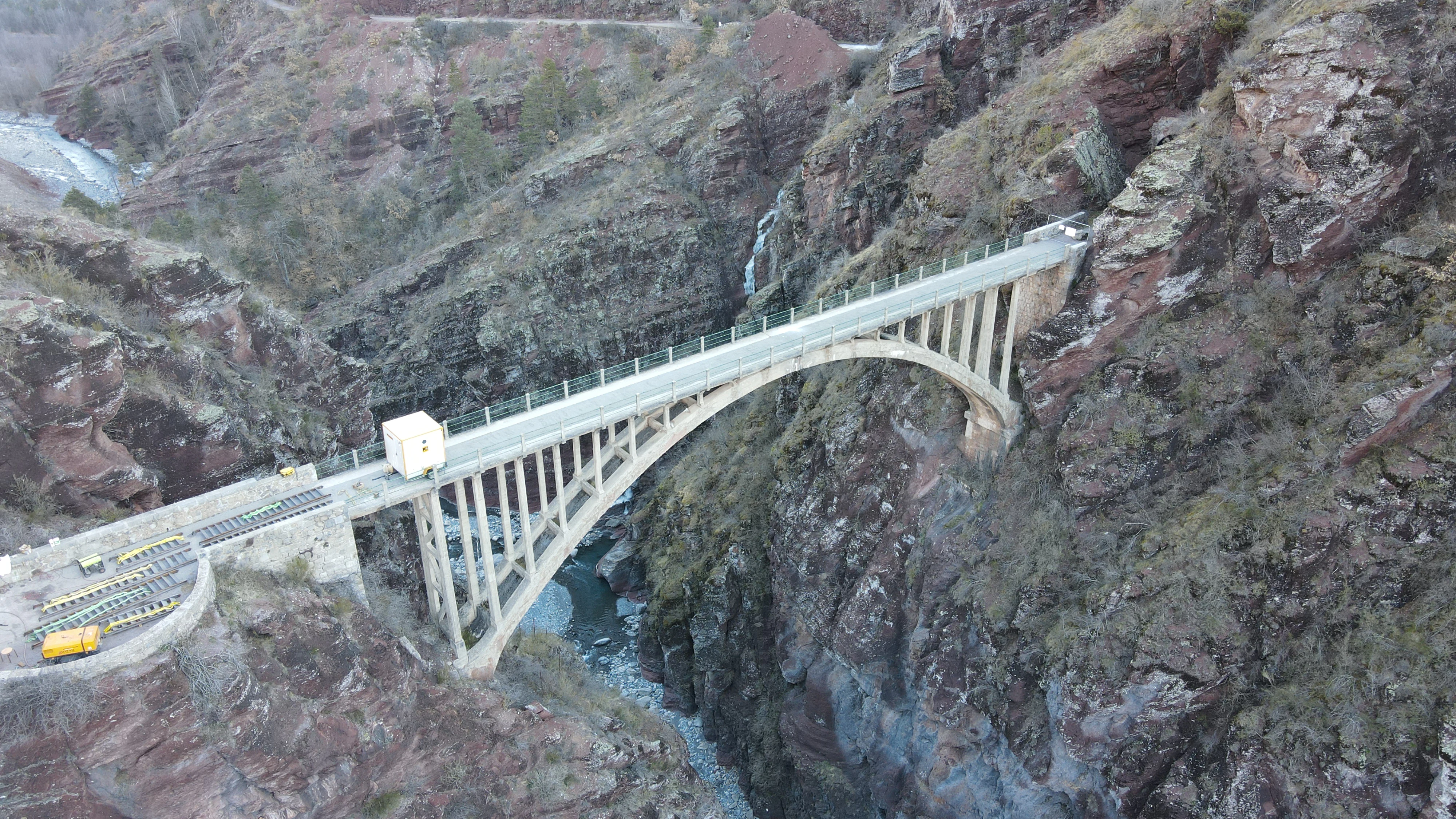 An aerial view of an arched concrete bridge, Pont de la Mariee, spanning a deep, narrow canyon with steep, reddish-brown rock walls and a river flowing below.