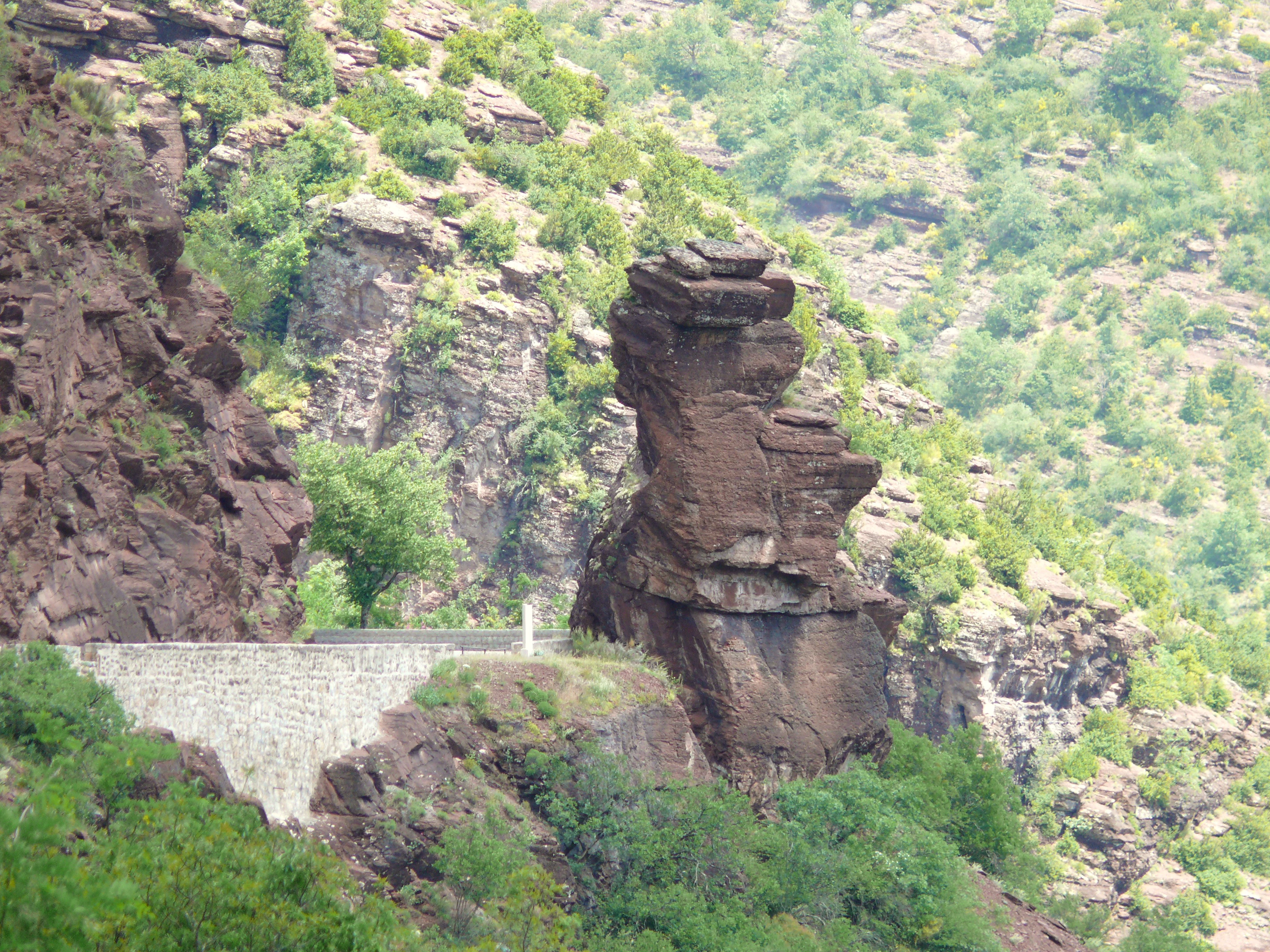 A distinctive red pelite rock formation known as Tete de Femme, set against a steep hillside with lush greenery and a stone retaining wall in the foreground.