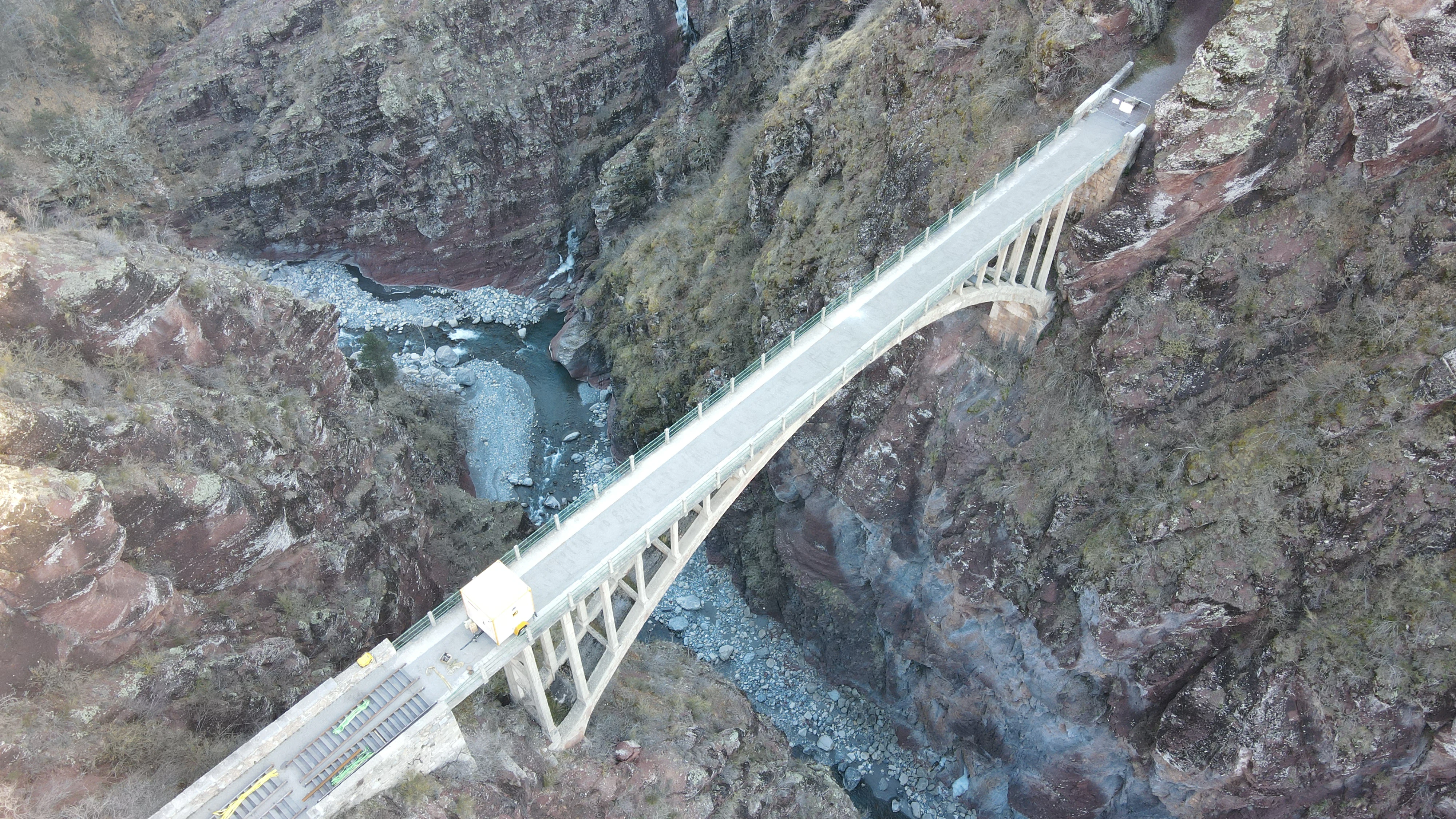 An aerial perspective of the arched Pont de la Marie bridge crossing a narrow, deep gorge with rugged red rock walls and a river flowing below.