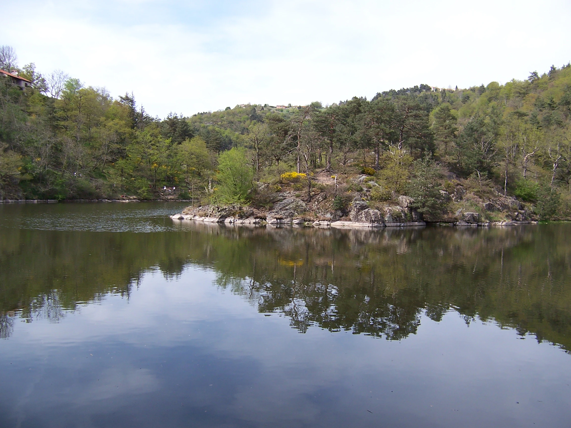 A peaceful lake surface reflecting a rocky, forested islet under a clear, bright sky.