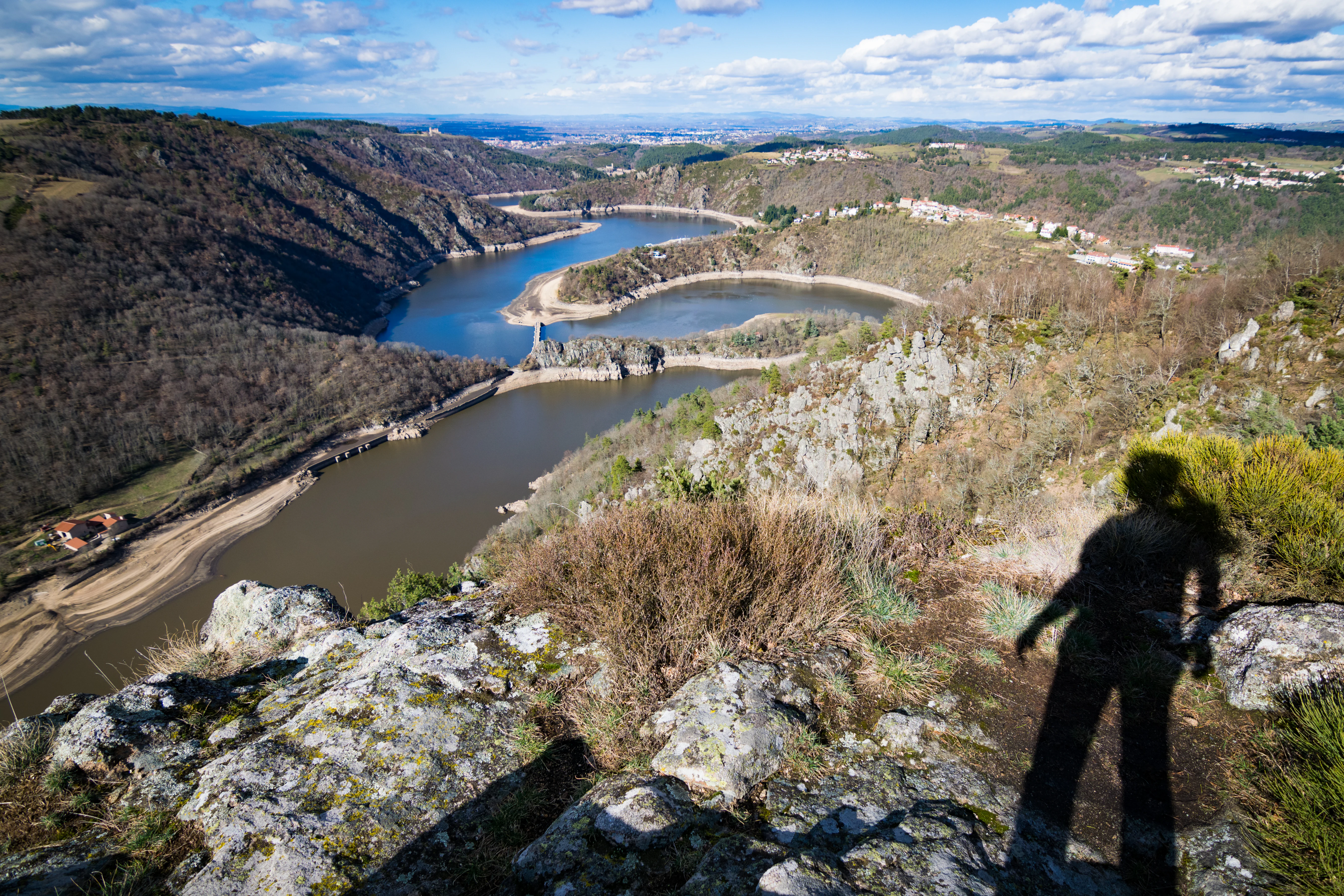 A high-angle scenic view looking down over a river that meanders through deep, forested gorges, with a rocky foreground and the shadow of a person visible on the right.