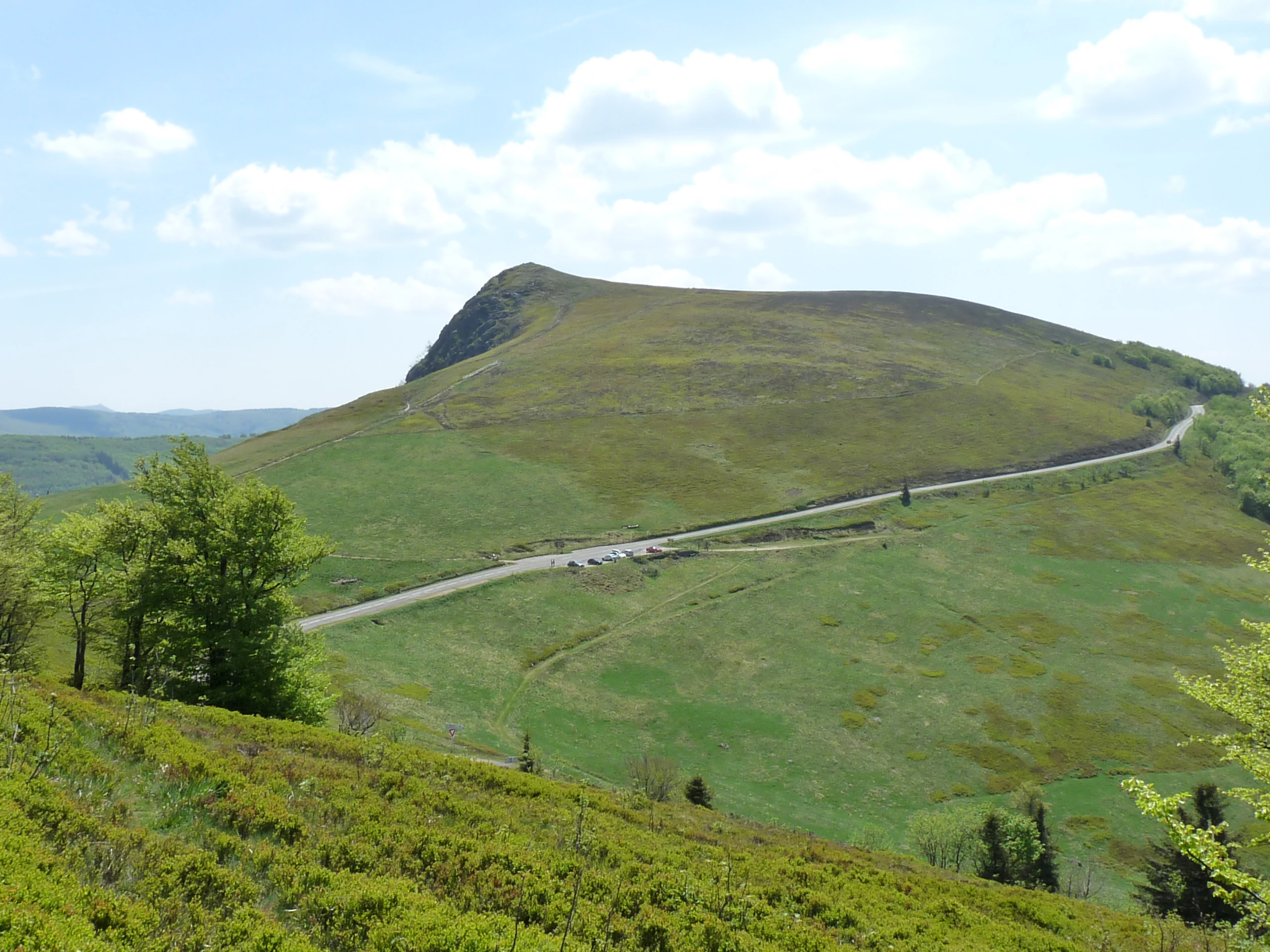 A wide shot of a rounded mountain peak covered in green grass, with a paved road curving along the slope and a small parking area in the distance.