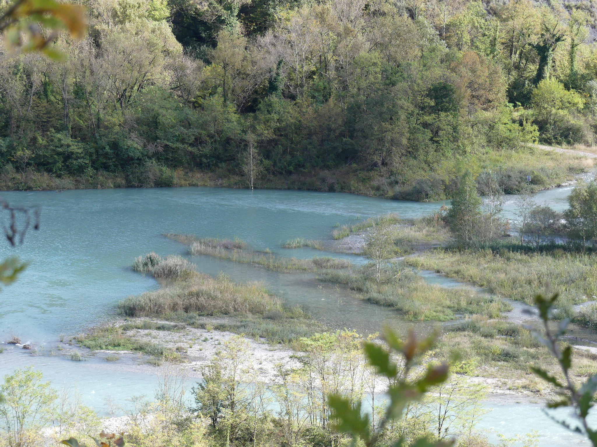 A wide view of a turquoise river winding through a wooded landscape, with low-lying islands, gravel banks, and riparian vegetation.