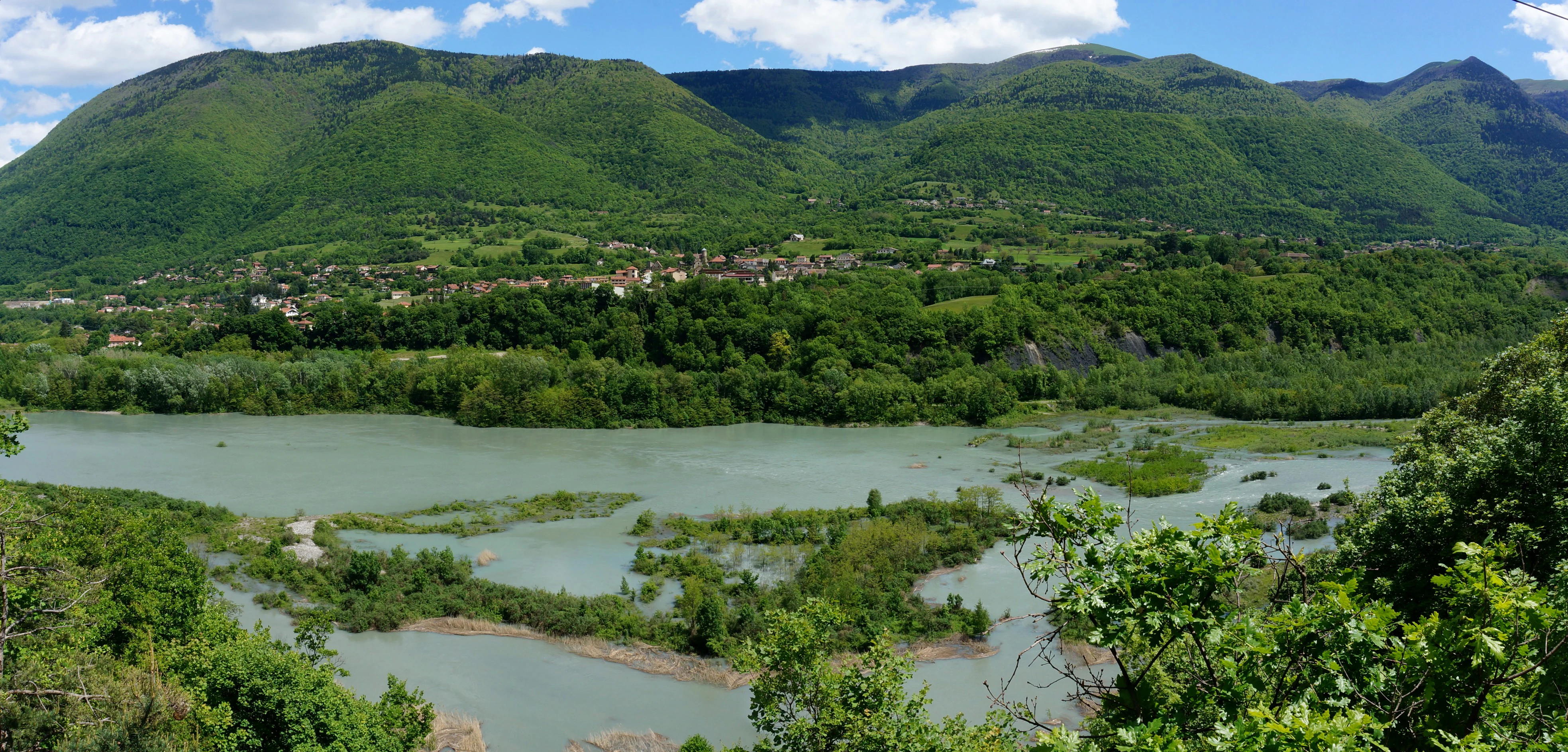 A wide view of the Drac River valley featuring a wide, silt-laden river with small islands and vegetation, set against a background of green, forested mountains under a blue sky.