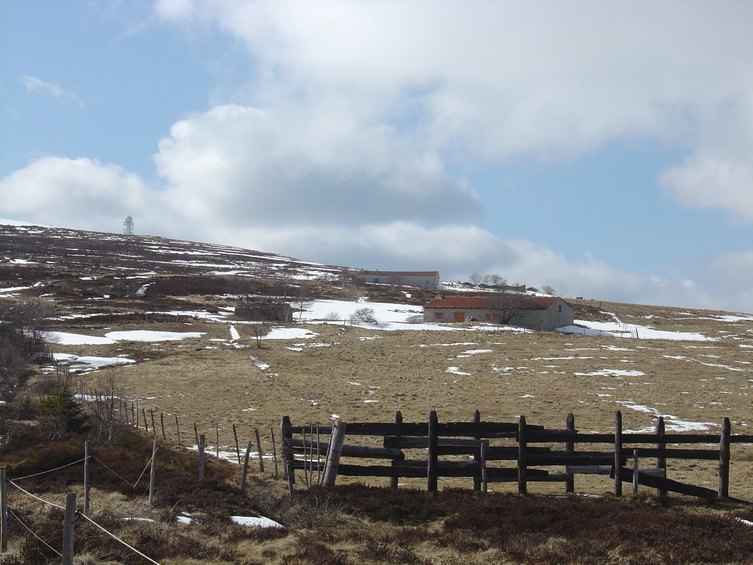 A wide view of a sloping, grassy mountain field under a cloudy sky, with patches of snow on the ground and several traditional low-slung stone buildings with reddish roofs in the mid-distance.